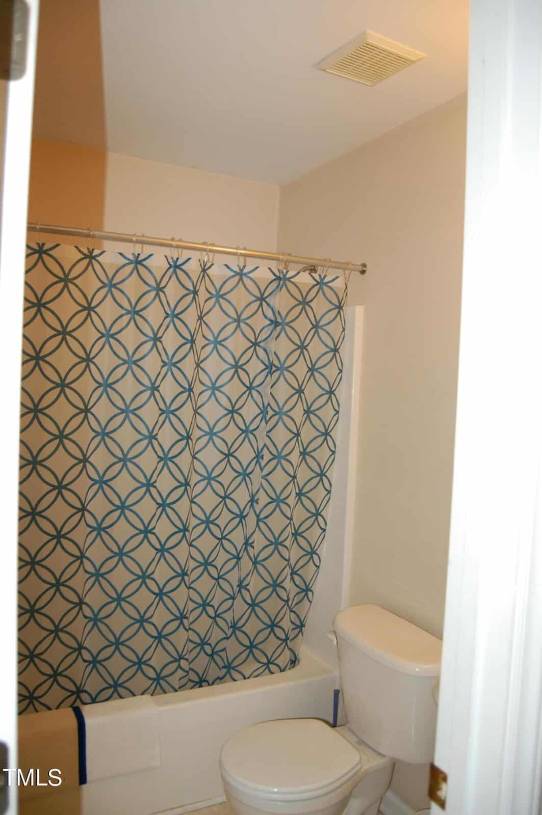 Bathroom with a shower-tub combo, featuring a blue and white geometric patterned curtain. A white toilet with the seat down is in the foreground. The walls are light-colored, and a small portion of a towel is visible hanging near the tub.