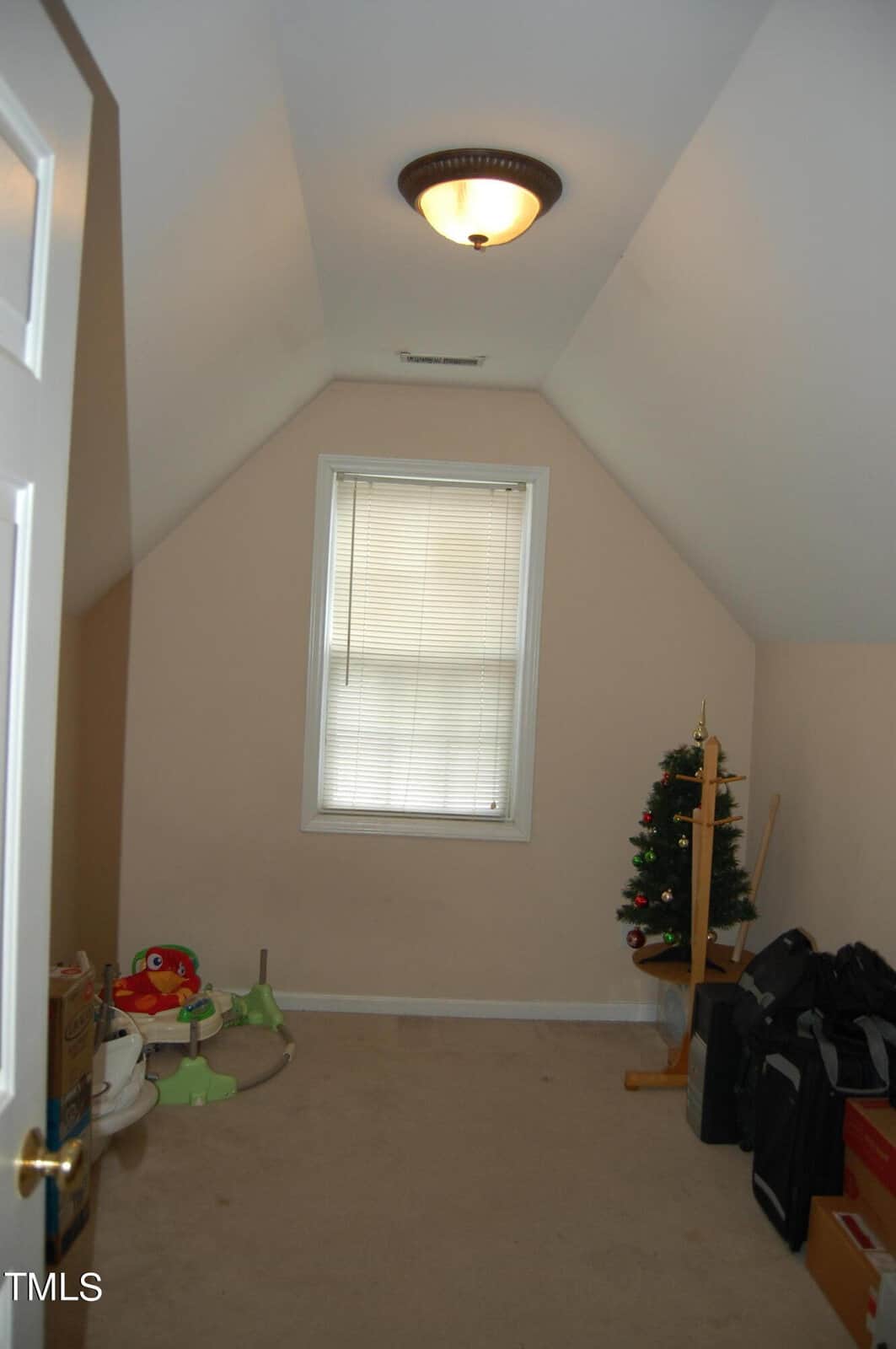 A small attic room with sloped ceilings, a single window with blinds, and a ceiling light. The room contains a small decorated Christmas tree, a few boxes, and some scattered children's toys.