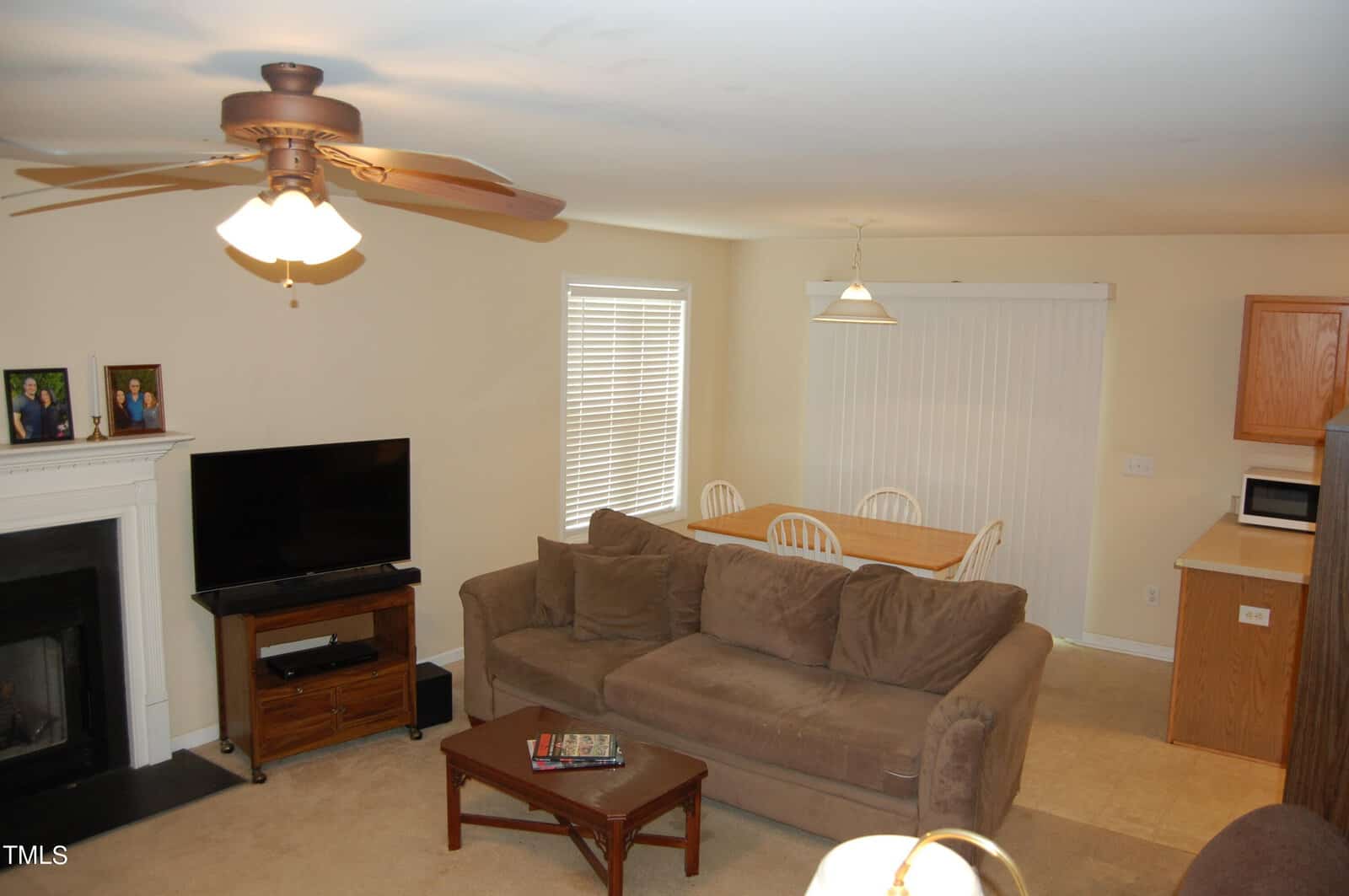A cozy living room features a brown sofa, wooden coffee table, and TV on a stand near a fireplace. A ceiling fan is above. In the background, there's a dining table with chairs and a kitchen area with a microwave. Family photos are displayed on the mantel.