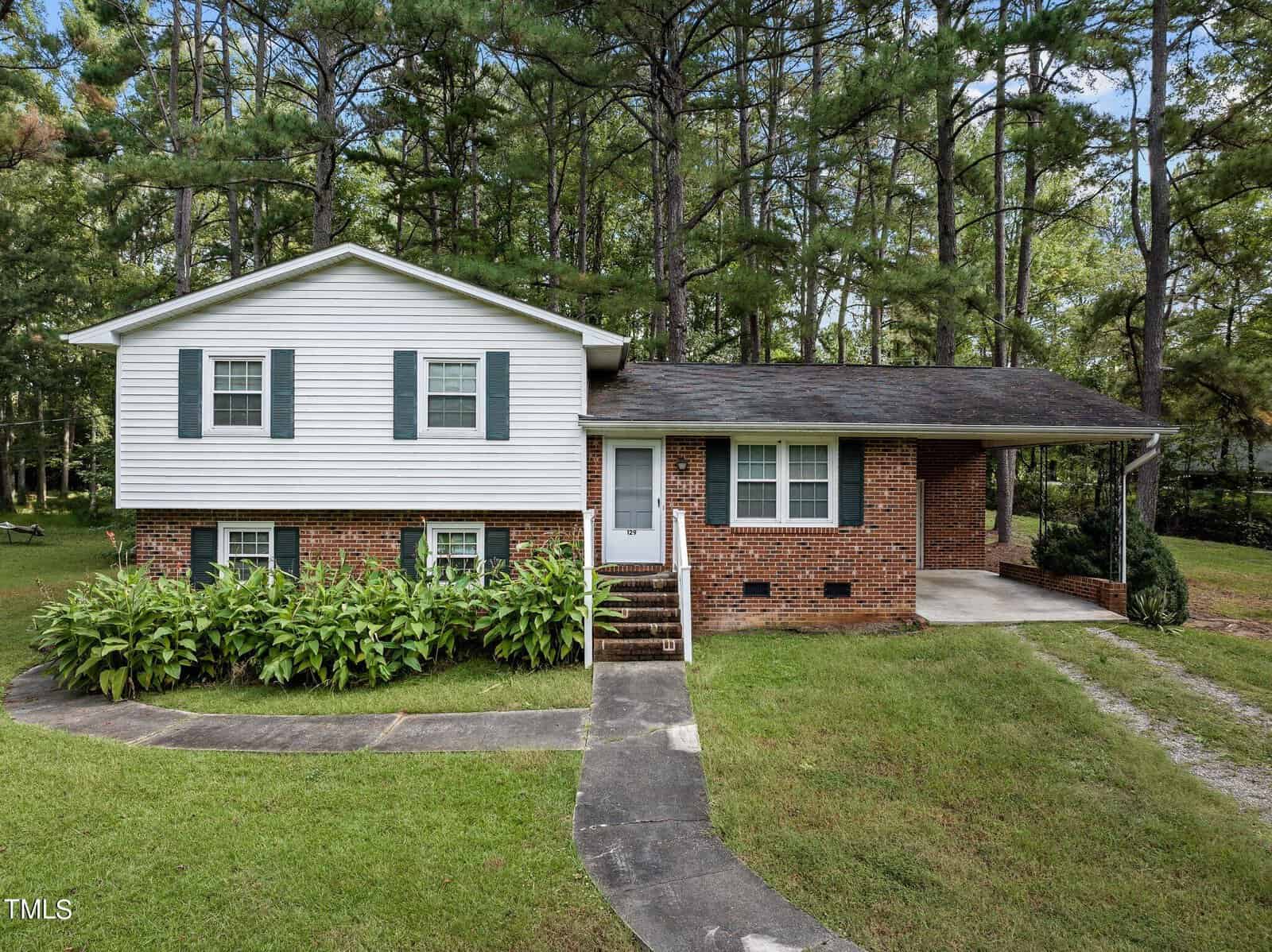 At 129 Maluli Drive in Oxford, this split-level house boasts a charming brick and white siding exterior, nestled among tall trees and bushes. A small set of stairs guides you to the front door, while a neatly cut lawn complements the driveway on the right.