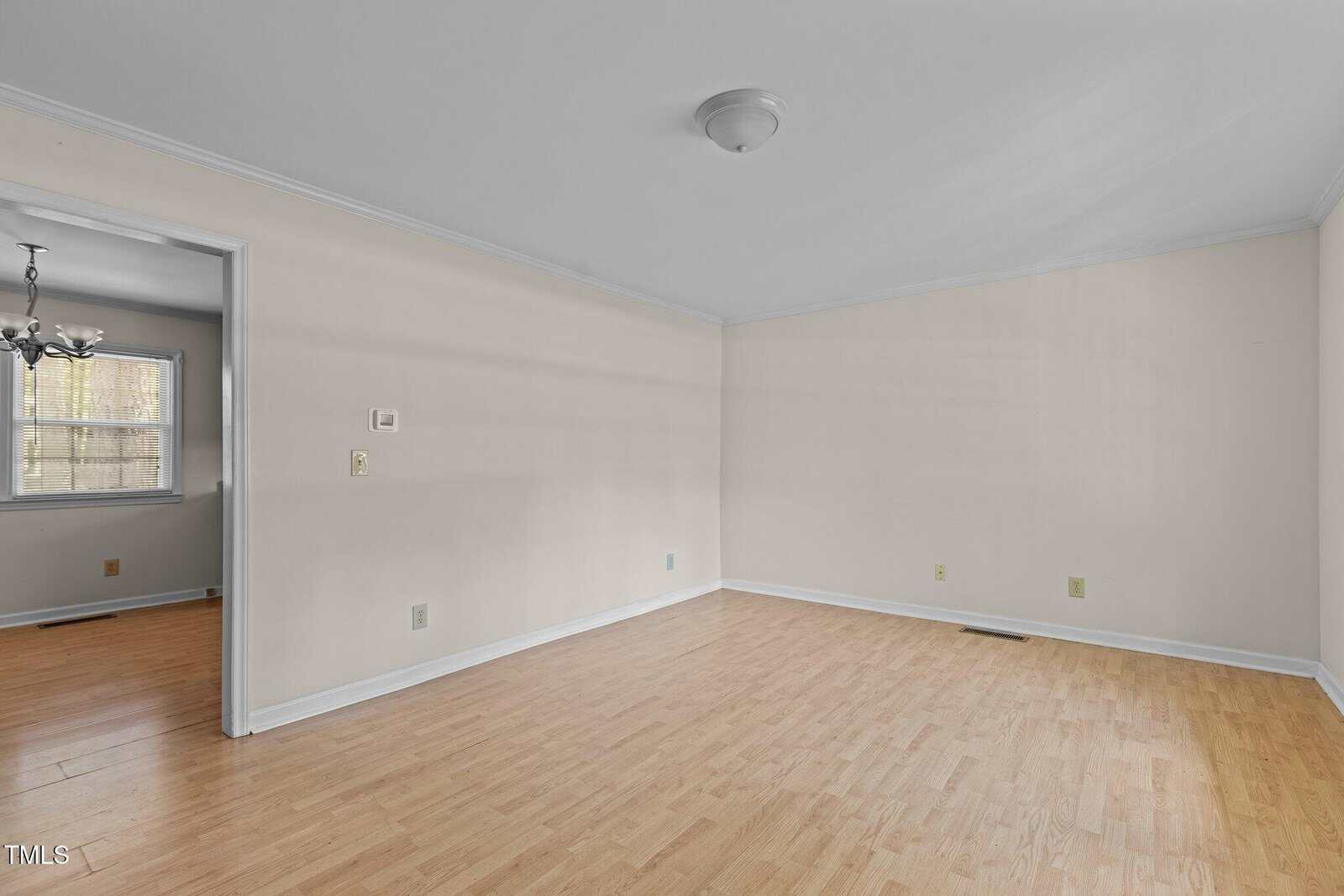 An empty room with light wood flooring and beige walls. A ceiling light fixture is centered in the room. An open doorway leads to another room with a window and a chandelier. Natural light enters through the windows.