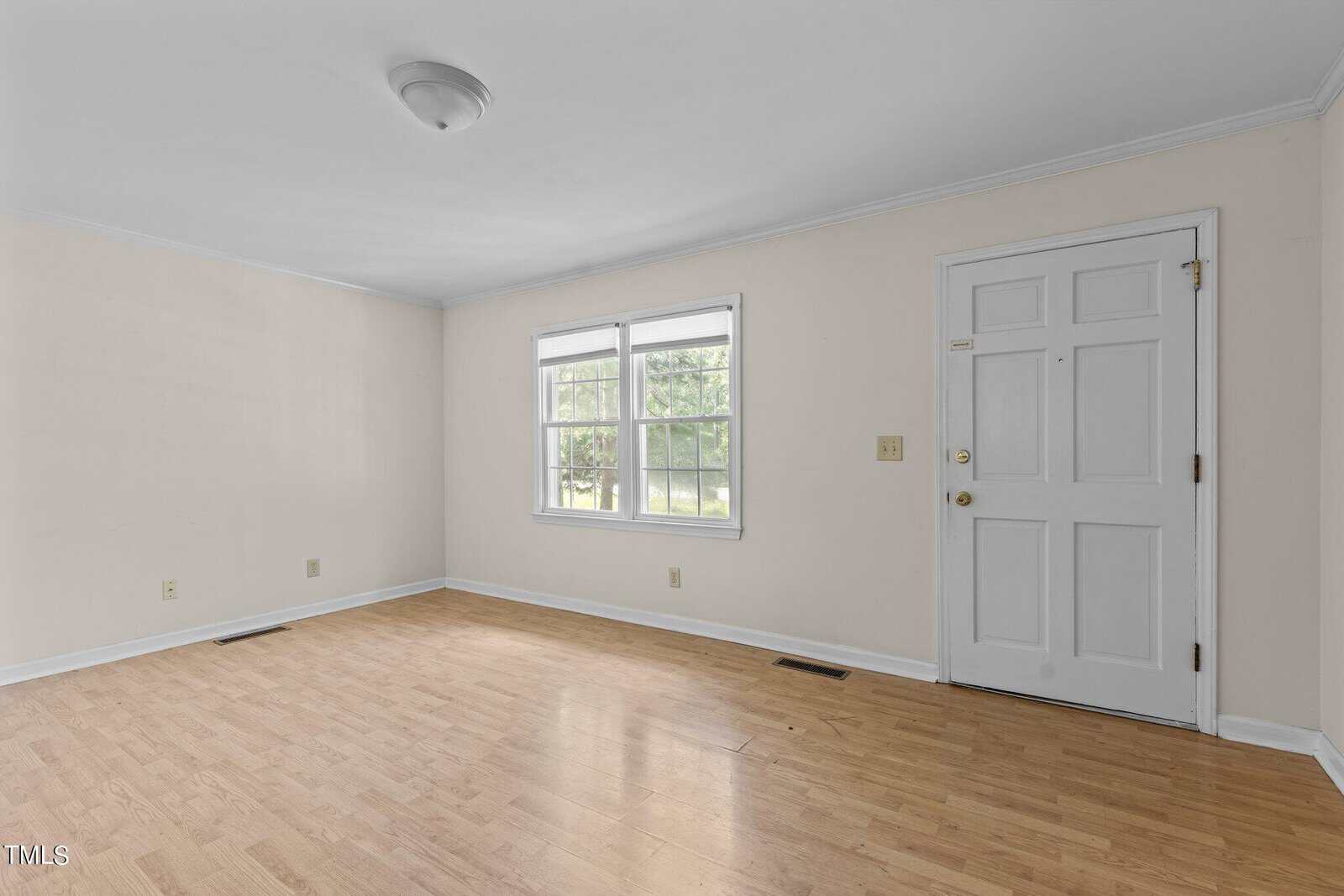An empty room with light wood flooring and cream-colored walls. It has a window with blinds and a white door. A ceiling light fixture is centered in the room, and there are electrical outlets on the walls.