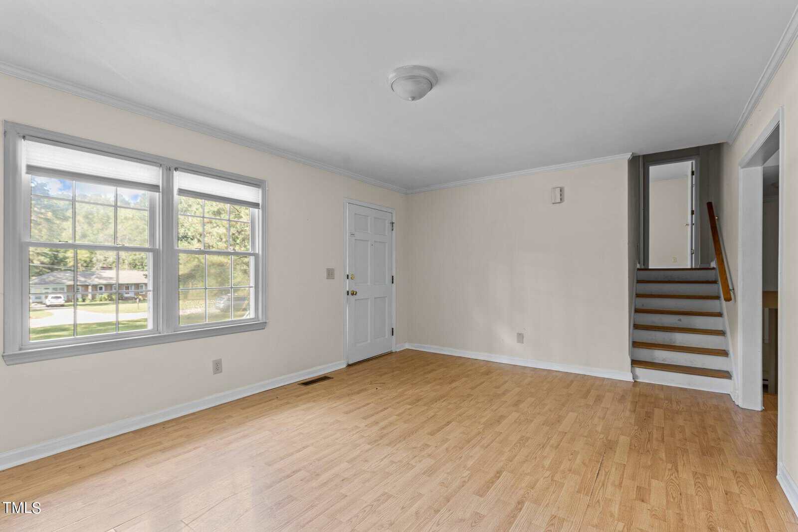 A bright, empty room with light wood flooring and off-white walls. It features a large window, a white door, and a staircase with a wooden handrail. Daylight illuminates the space, and the ceiling has a simple light fixture.