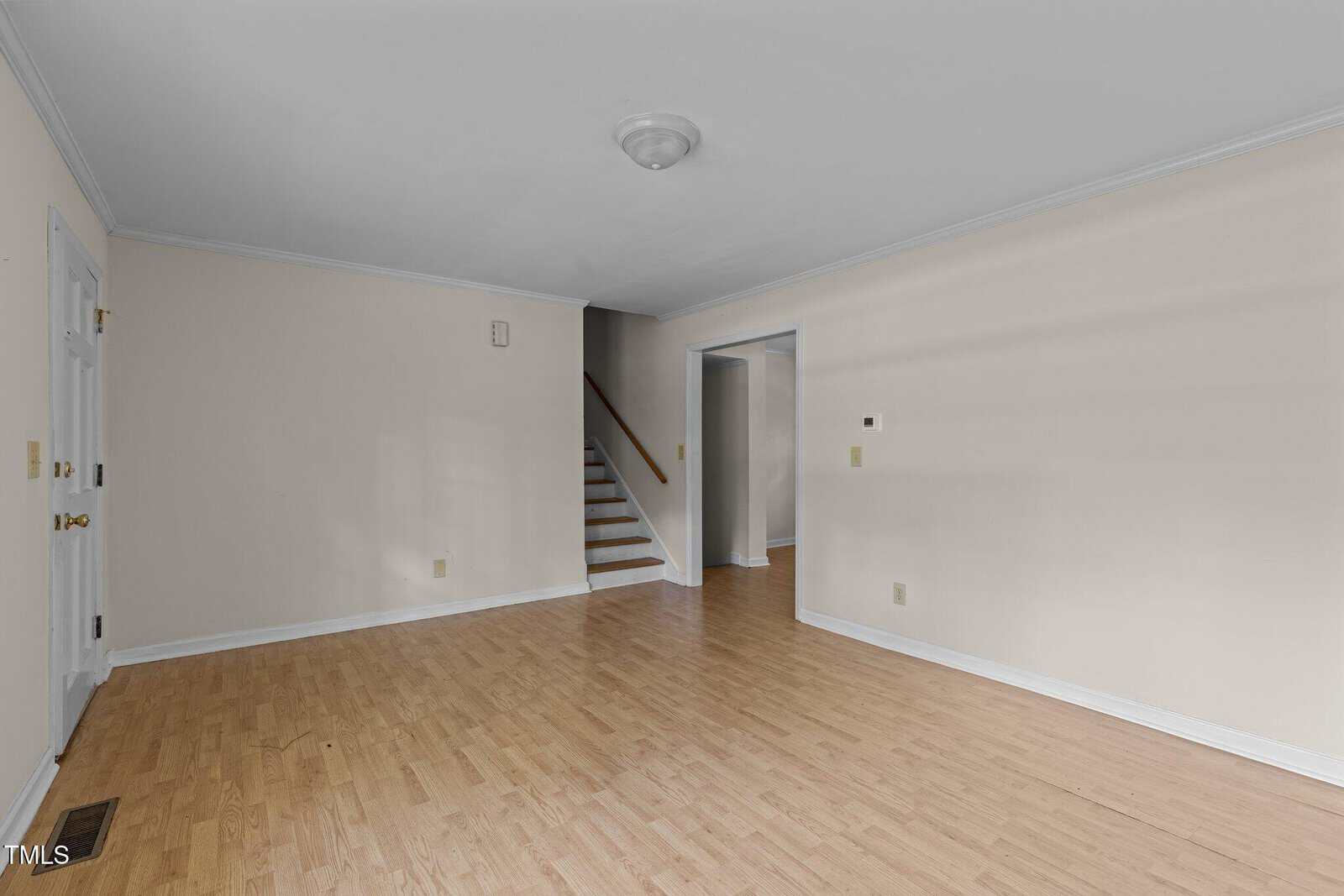 An empty room with light-colored walls and wooden flooring. There is a staircase leading upward, a closed white door, and a small ceiling light fixture. The room is well-lit with natural light from an unseen source.