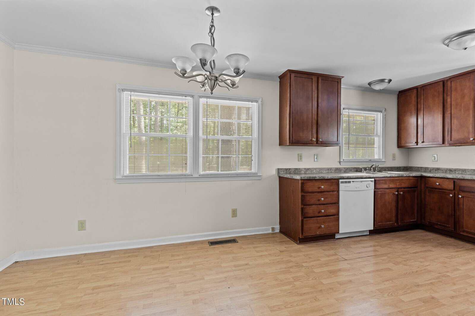 A kitchen with wooden cabinets, a marble countertop, and a stainless steel dishwasher. There's a two-window setup with blinds, a double sink, and a ceiling-mounted light fixture. The floor has a wooden finish, and the walls are light-colored.