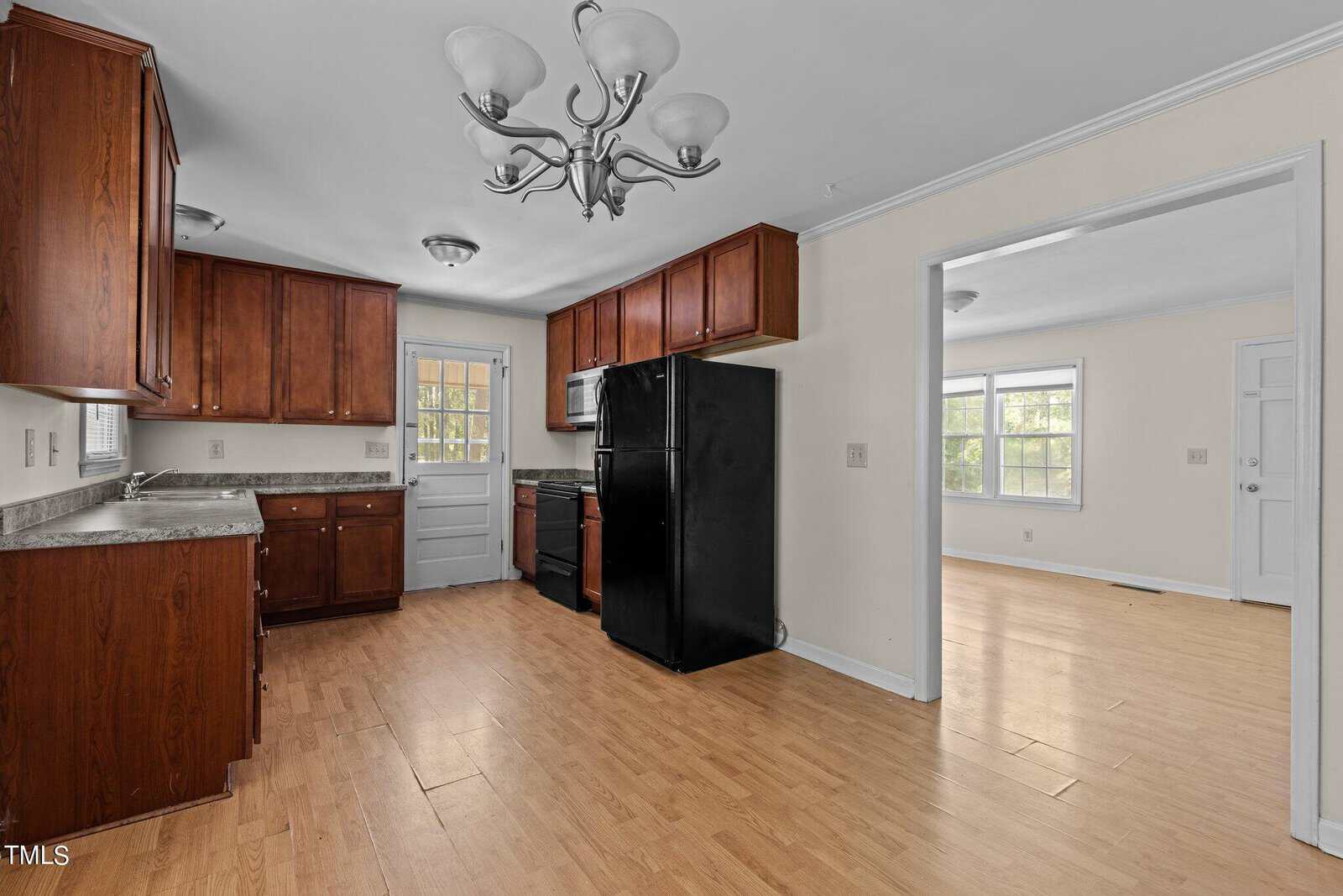 A kitchen with light wooden flooring, brown wooden cabinets, and a black fridge. The room includes a black stove and a window above the countertop. A light fixture hangs from the ceiling, and an adjoining room is visible through an open doorway.