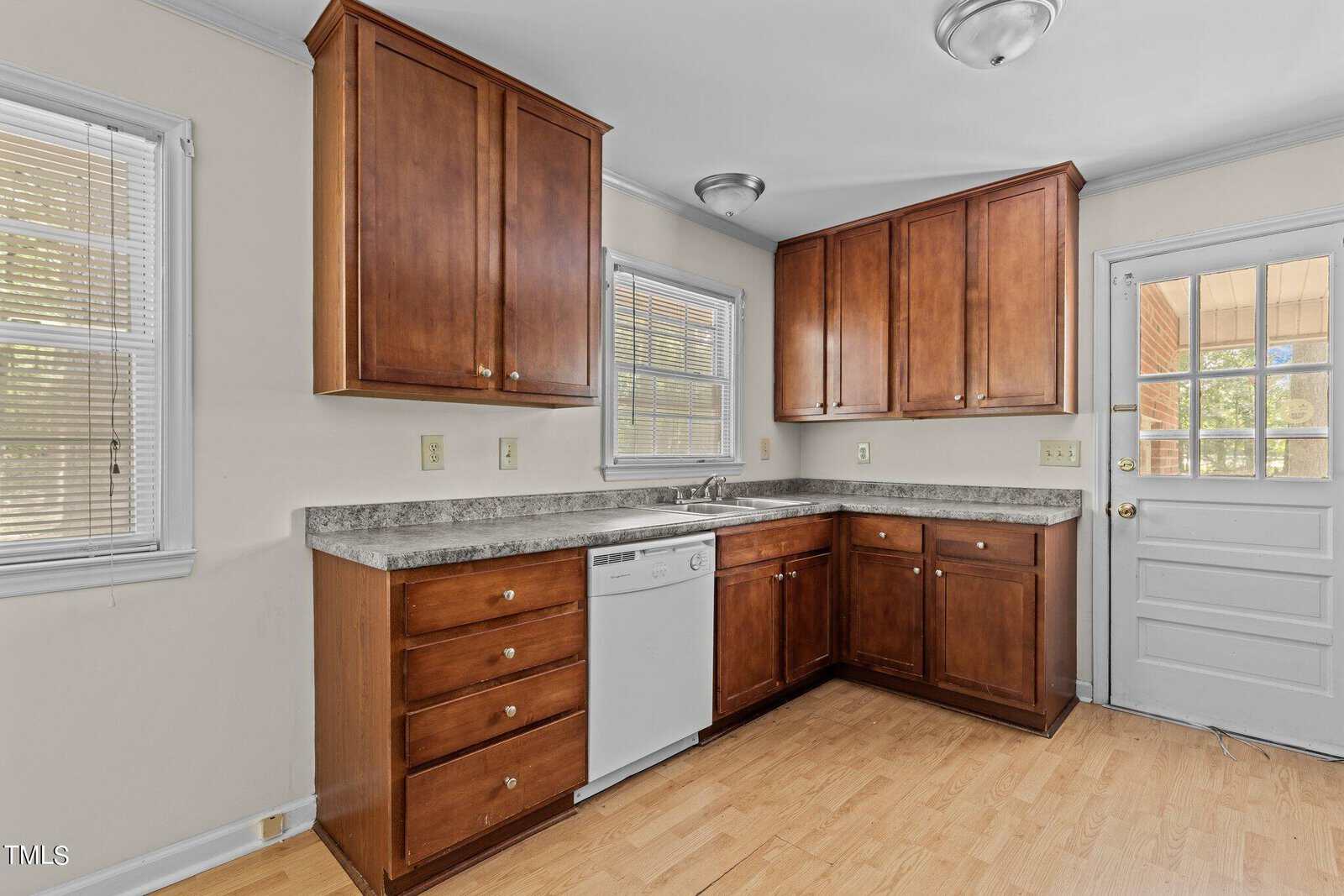 A kitchen with wooden cabinets, a dishwasher, and granite countertops. There are two windows and a door with glass panes leading outside. The floor is light wood, and the walls are light-colored. Ceiling lights are visible.