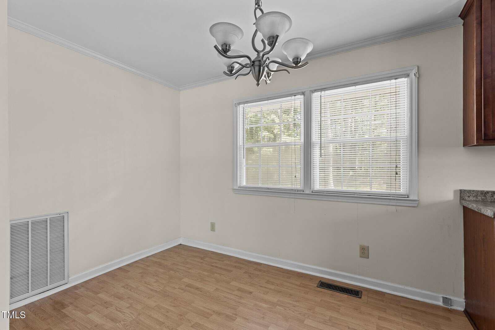 An empty room with light-colored walls and wooden flooring. There is a three-light chandelier on the ceiling. Two windows with blinds allow natural light in. A vent is on the lower wall next to a wooden cabinet on the right.