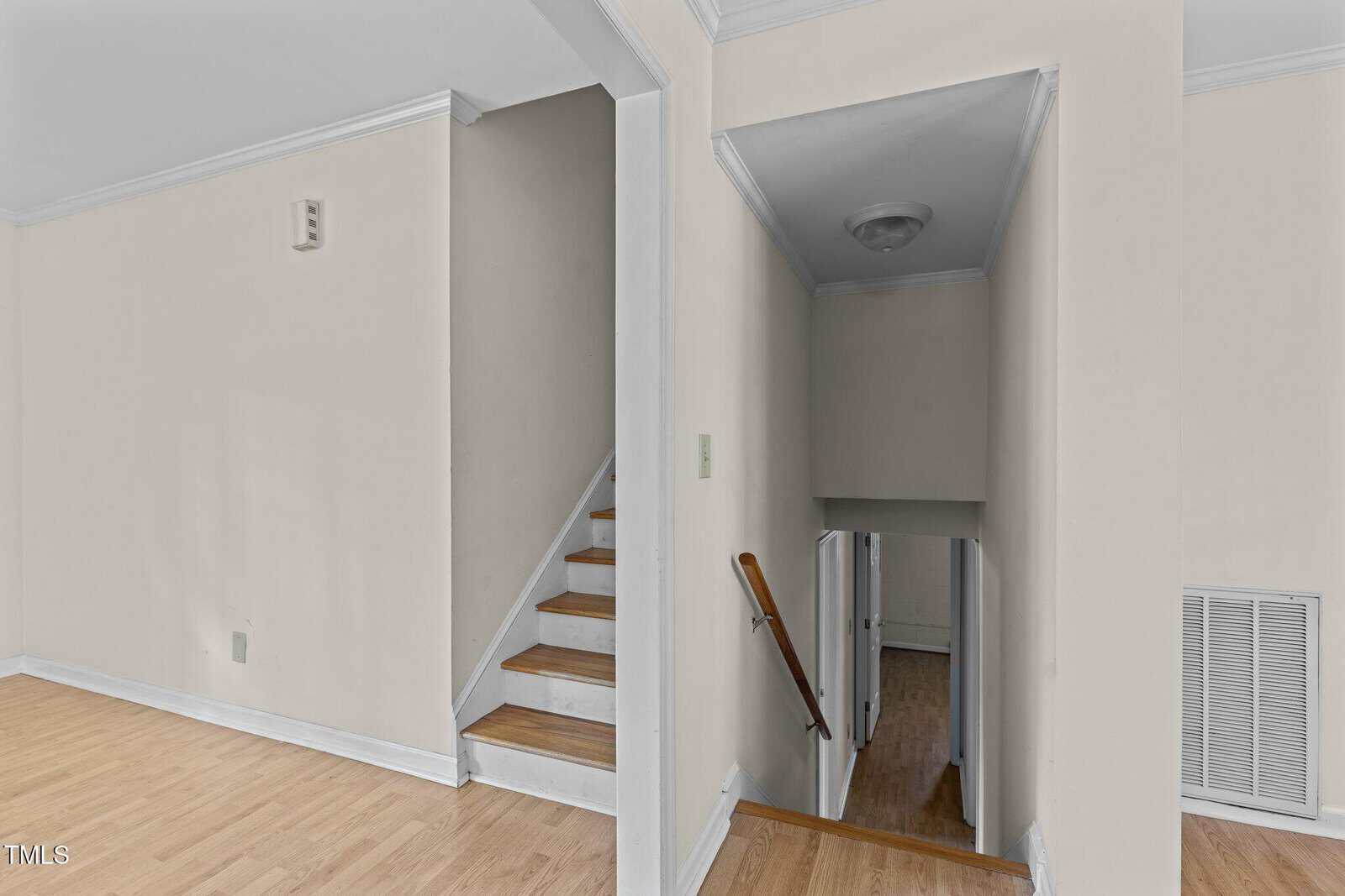 A view of an indoor staircase area in a home. The image shows a set of wooden stairs ascending to the left with a white handrail. To the right, there is a hallway with light wood flooring, leading to a door at the end. Walls are painted light beige.