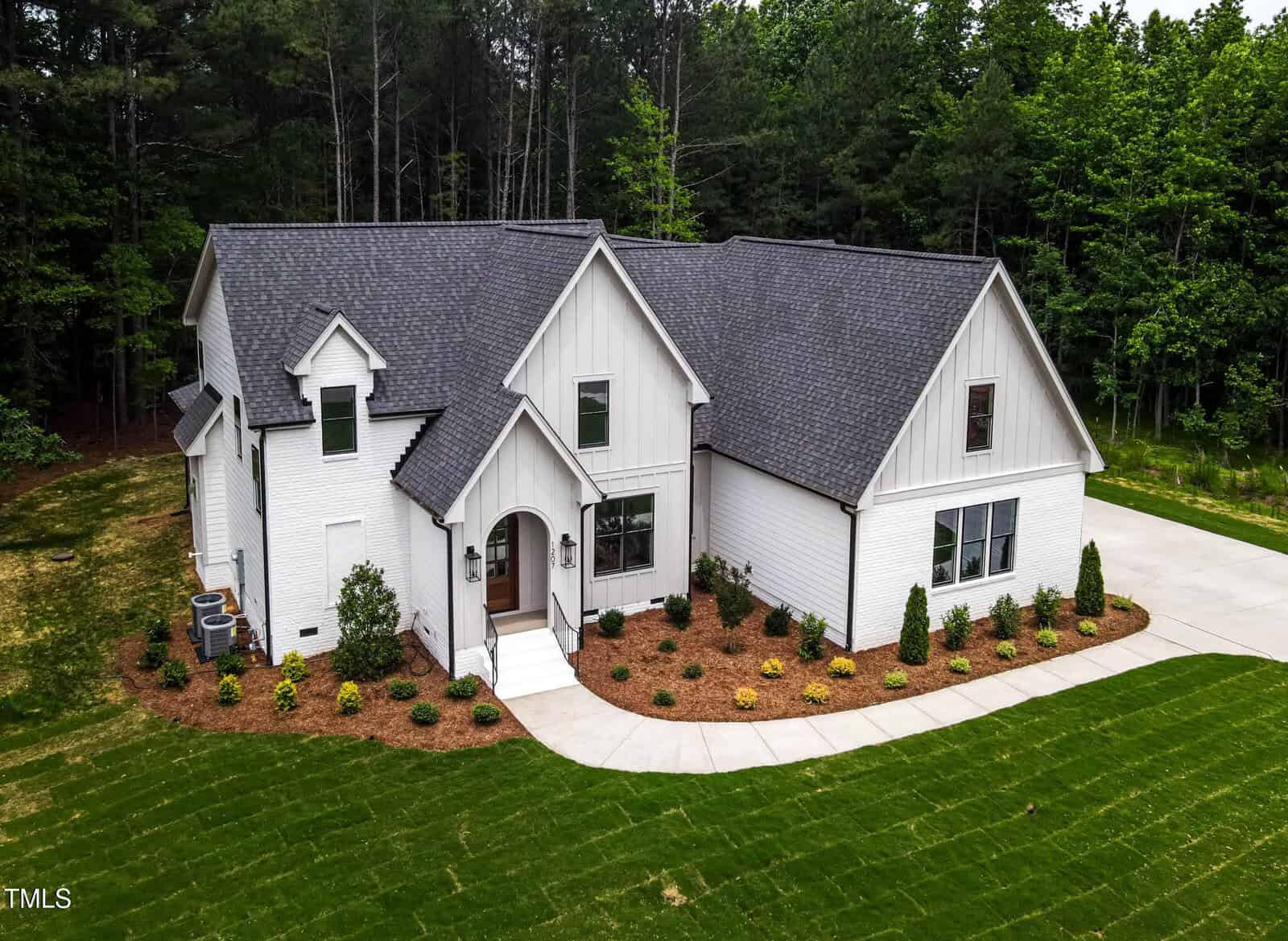 A modern white brick house with dark gray roof shingles sits on a manicured lawn at 1207 Red Cedar Court, Youngsville, surrounded by landscaped bushes and trees, with a forested area behind and a concrete driveway to the right.