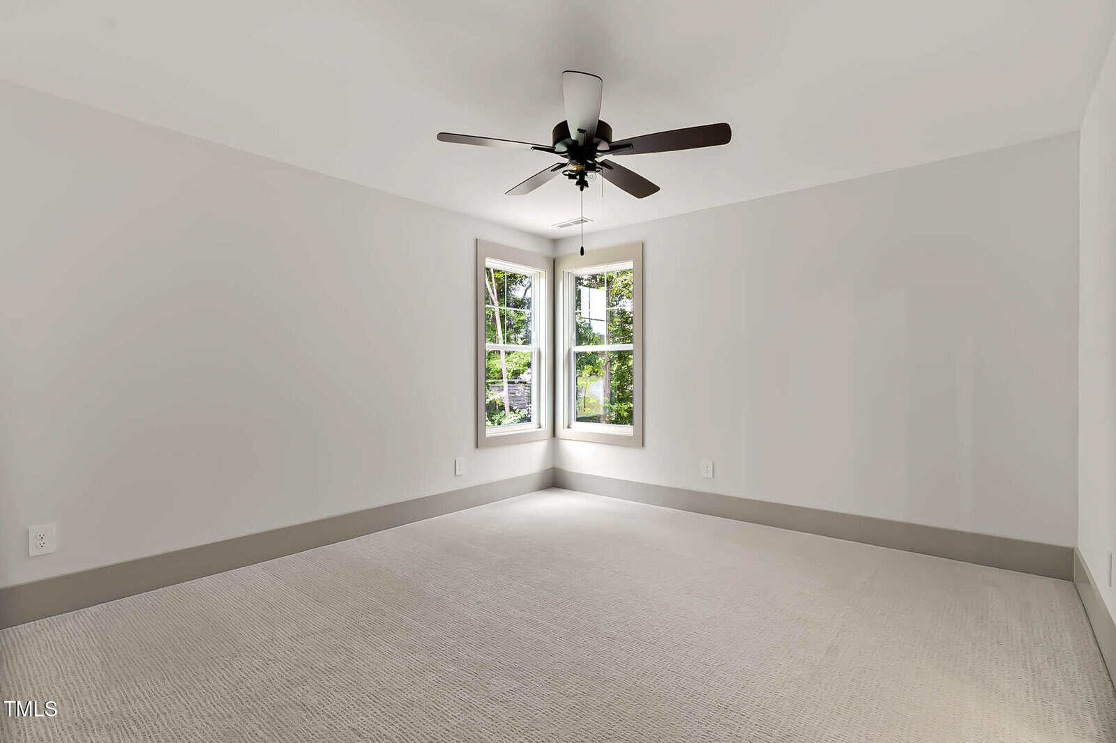 Bright, empty room with light-colored carpet, white walls, a ceiling fan with dark blades, and a pair of windows letting in natural light and showing greenery outside.