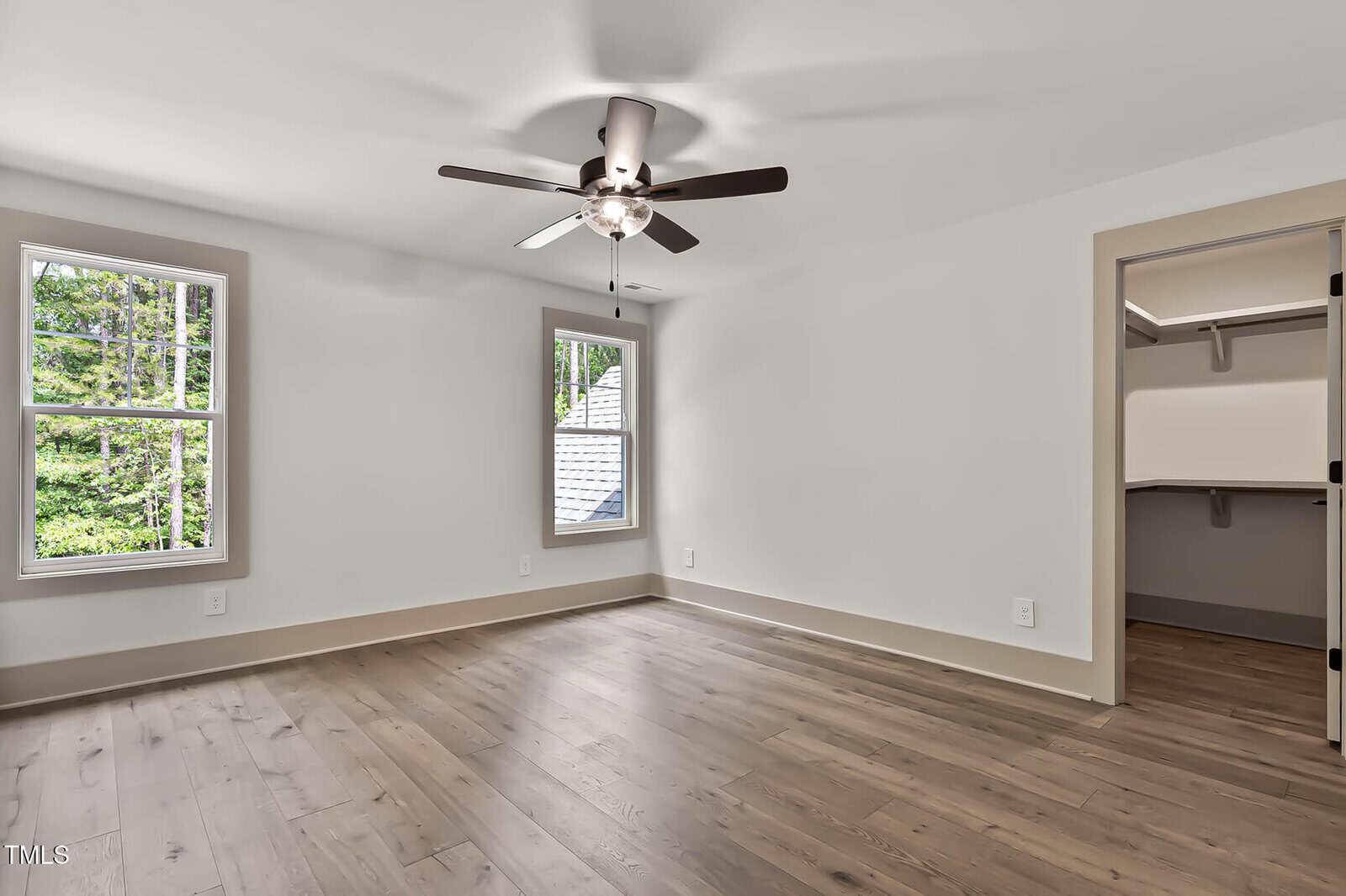 A bright, empty room with light wood flooring, white walls, two large windows, a ceiling fan with lights, and a doorway leading to a walk-in closet. Trees are visible through the windows.
