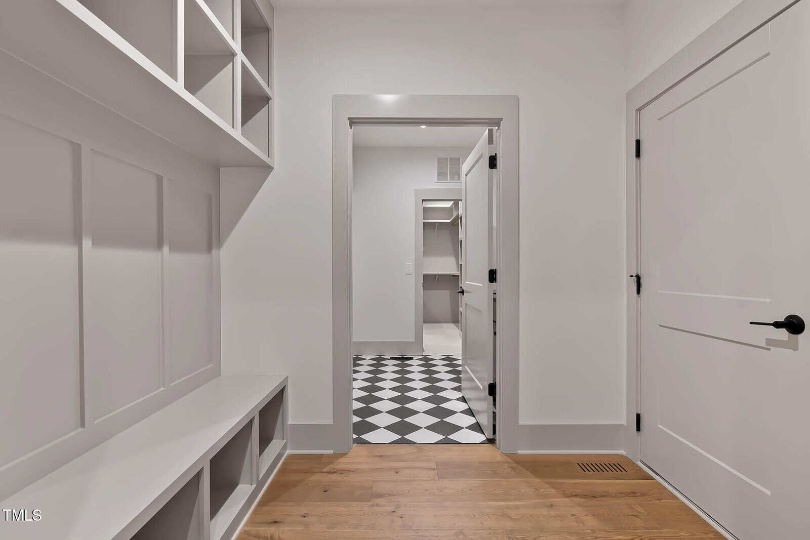 A modern hallway with white walls, built-in shelves and cubbies, light wood flooring, and black-and-white checkered tile flooring visible in the adjoining room. Two closed doors are on the right.