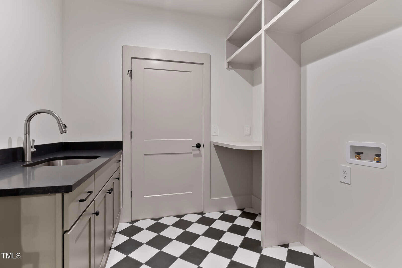 A modern laundry room with black and white checkered tile flooring, light gray cabinets, a black countertop with a sink, open shelving, and hookups for a washer and dryer.