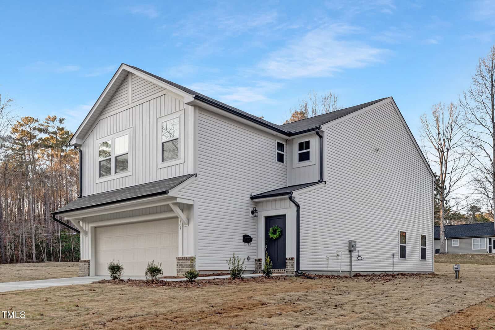 Two-story modern suburban house with light-colored vinyl siding, an attached two-car garage, black roof, and small shrubs in front. The house is situated on a large grassy lot with trees in the background.