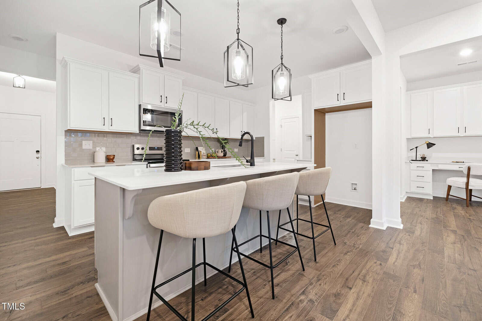 Modern kitchen with white cabinets, stainless steel appliances, a large island with three beige barstools, pendant lighting, wood flooring, and a built-in desk area in the background.