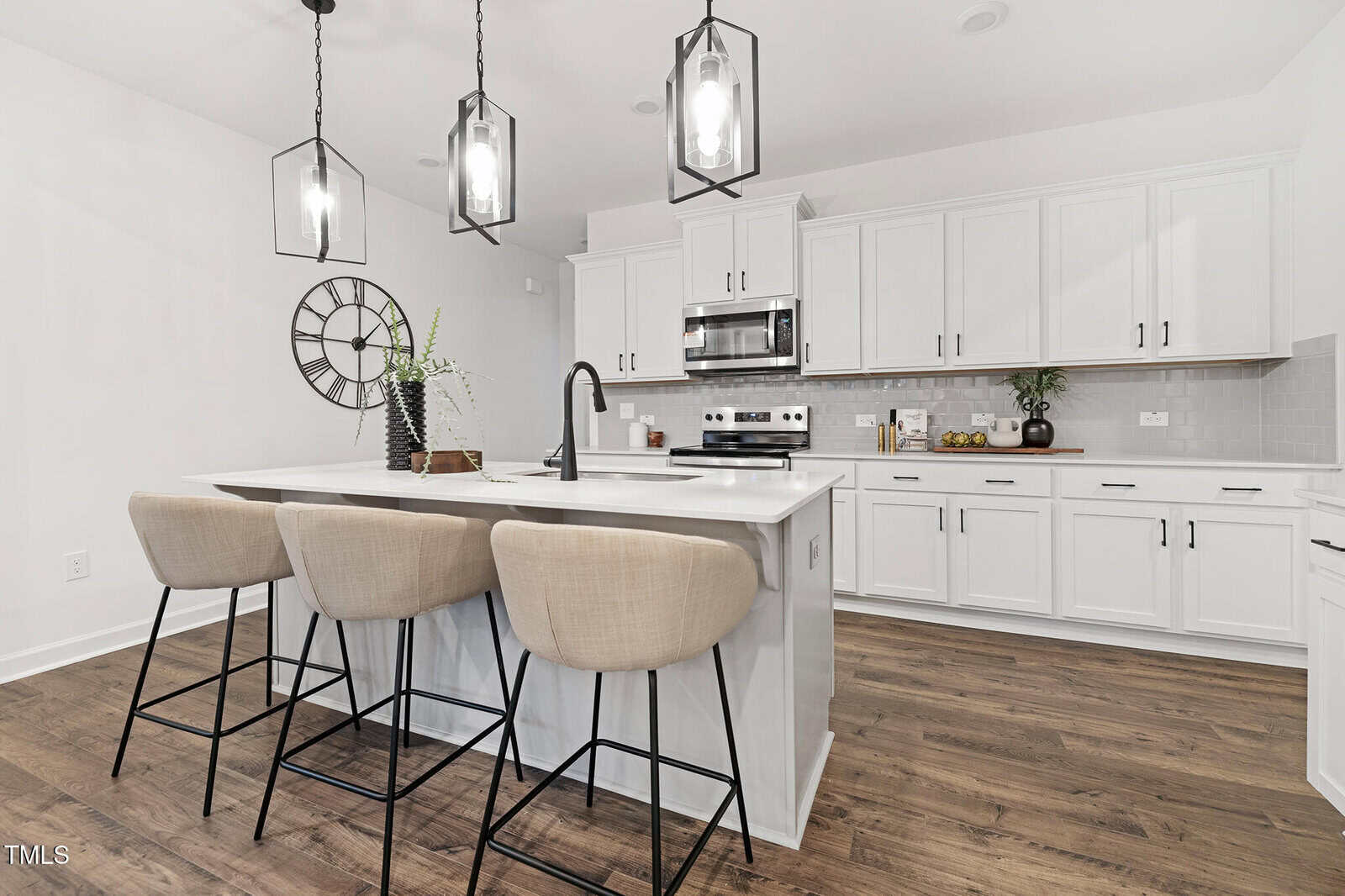 A modern kitchen with white cabinets, stainless steel appliances, an island with a sink and faucet, three beige barstools, pendant lights above the island, and wood flooring. A decorative clock hangs on the wall.