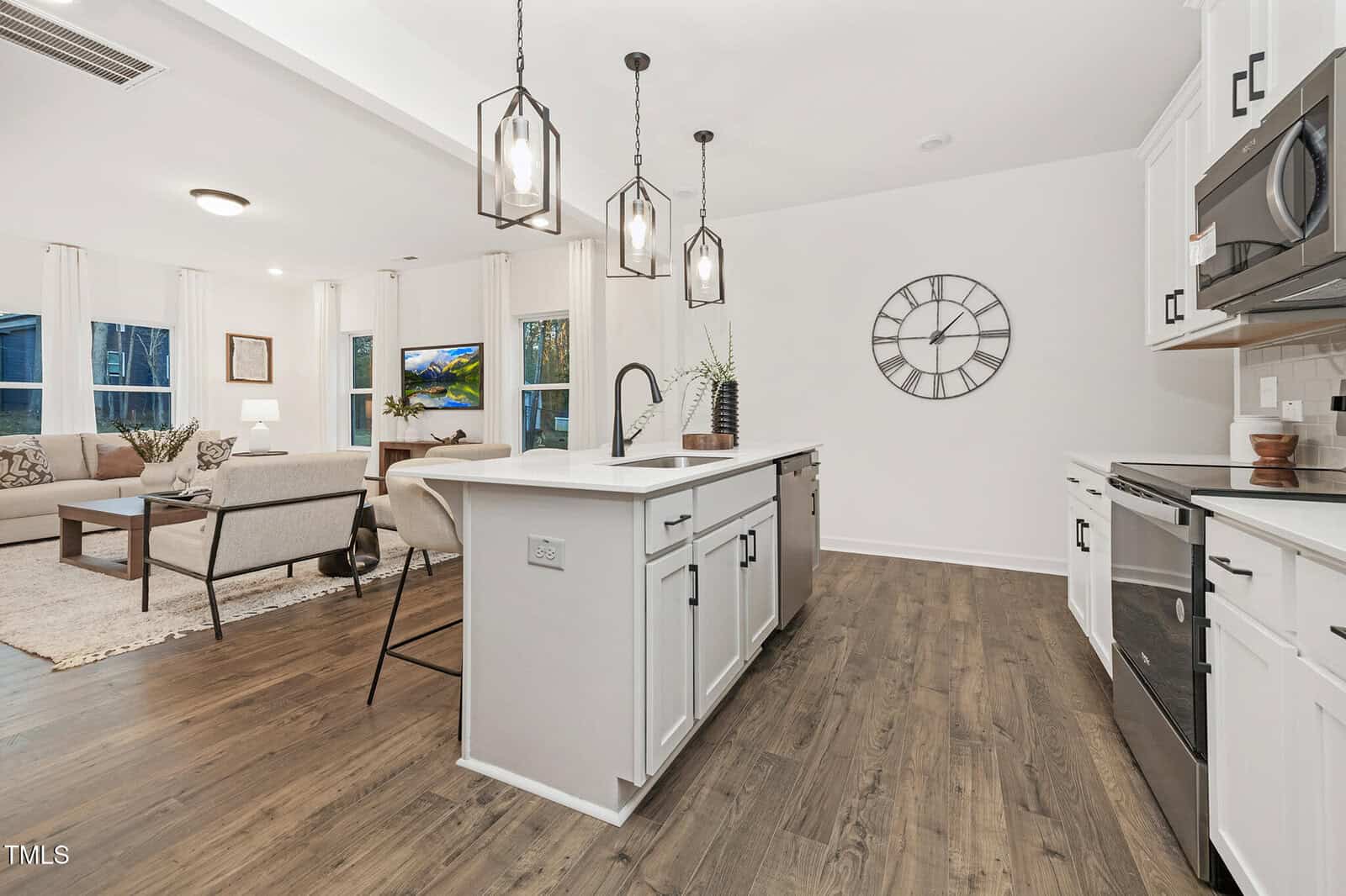 Modern open-concept kitchen and living room with white cabinets, a large island with a sink, pendant lights, a wall clock, wood flooring, and a seating area with a sofa, chairs, and a TV in the background.