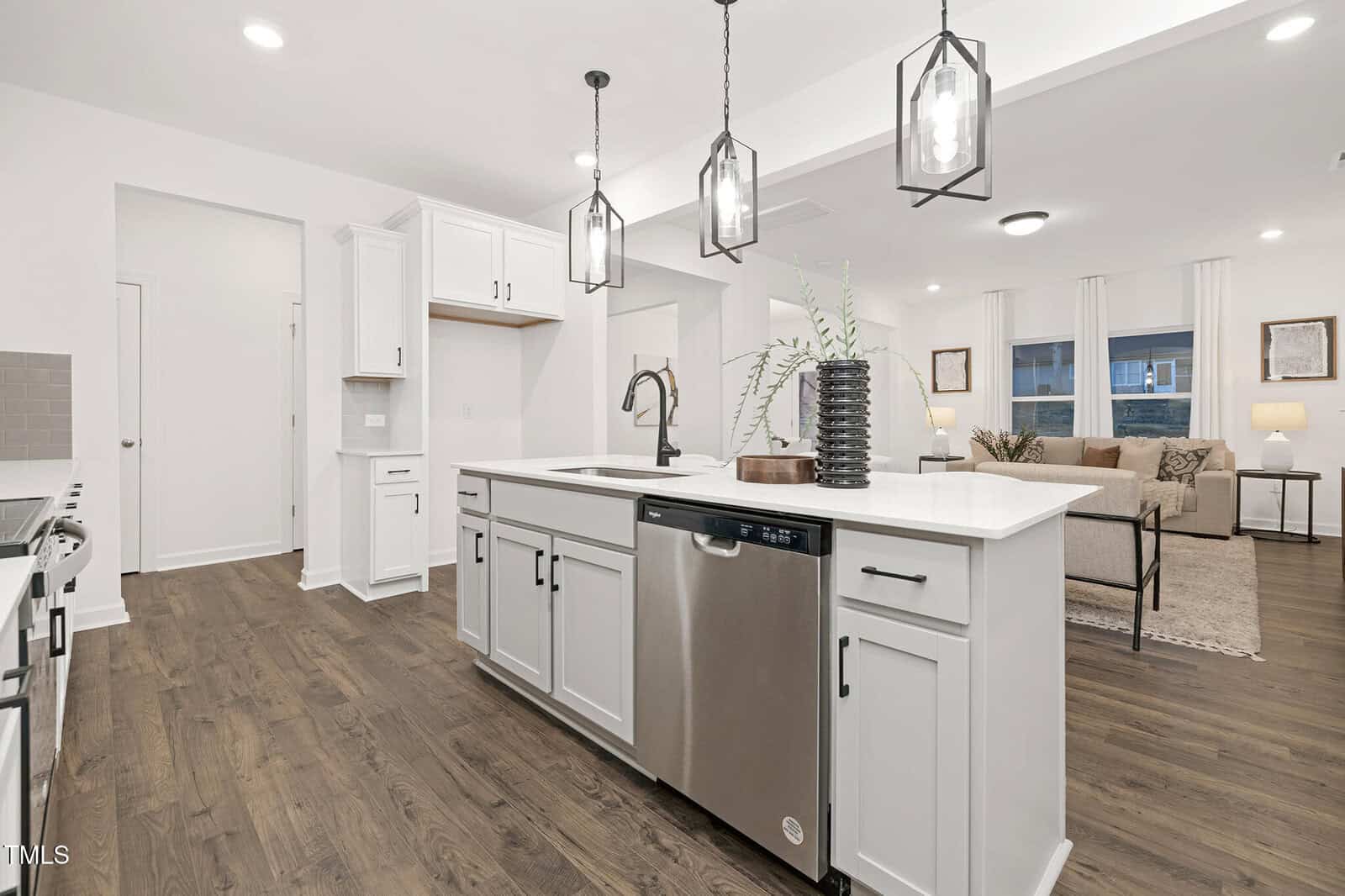 Modern kitchen with white cabinets, stainless steel appliances, and a large island with a sink and dishwasher. There are pendant lights above the island and a living area with a sofa and windows in the background.
