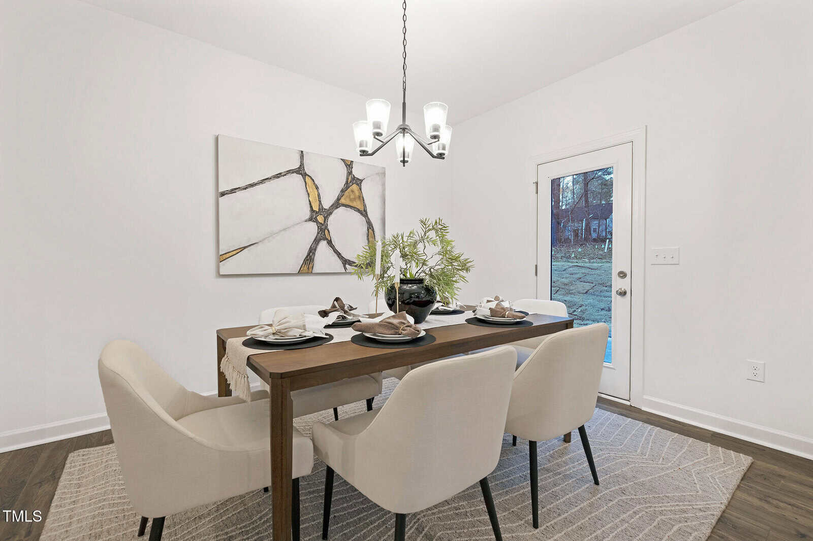 A dining room with a rectangular wooden table set for four, beige upholstered chairs, a light gray rug, abstract wall art, a modern chandelier, and a glass door leading outside.