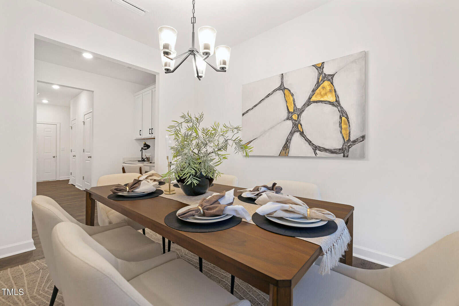 A modern dining room with a wooden table set for four, cream upholstered chairs, a potted plant centerpiece, neutral napkins, and a contemporary abstract painting with yellow and black accents on the wall.