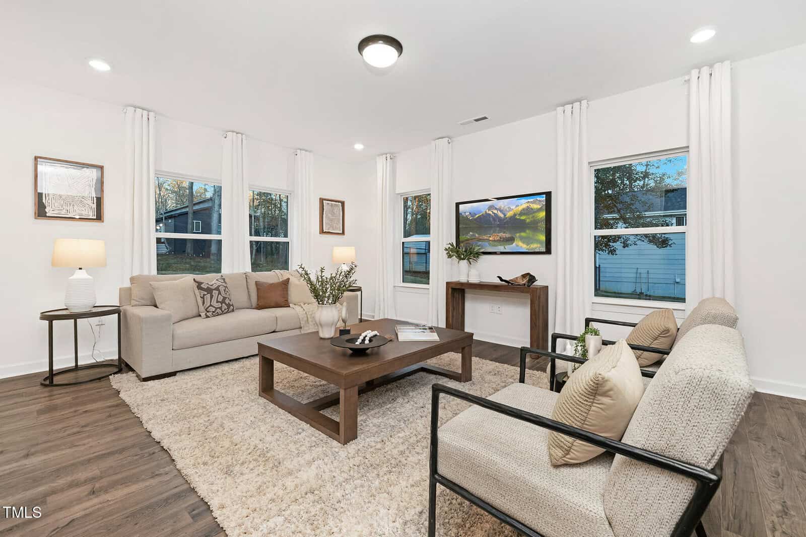 A modern living room with a beige sofa, two chairs, a wooden coffee table on a shag rug, wall art, table lamps, and a TV mounted above a console against white walls and large windows.
