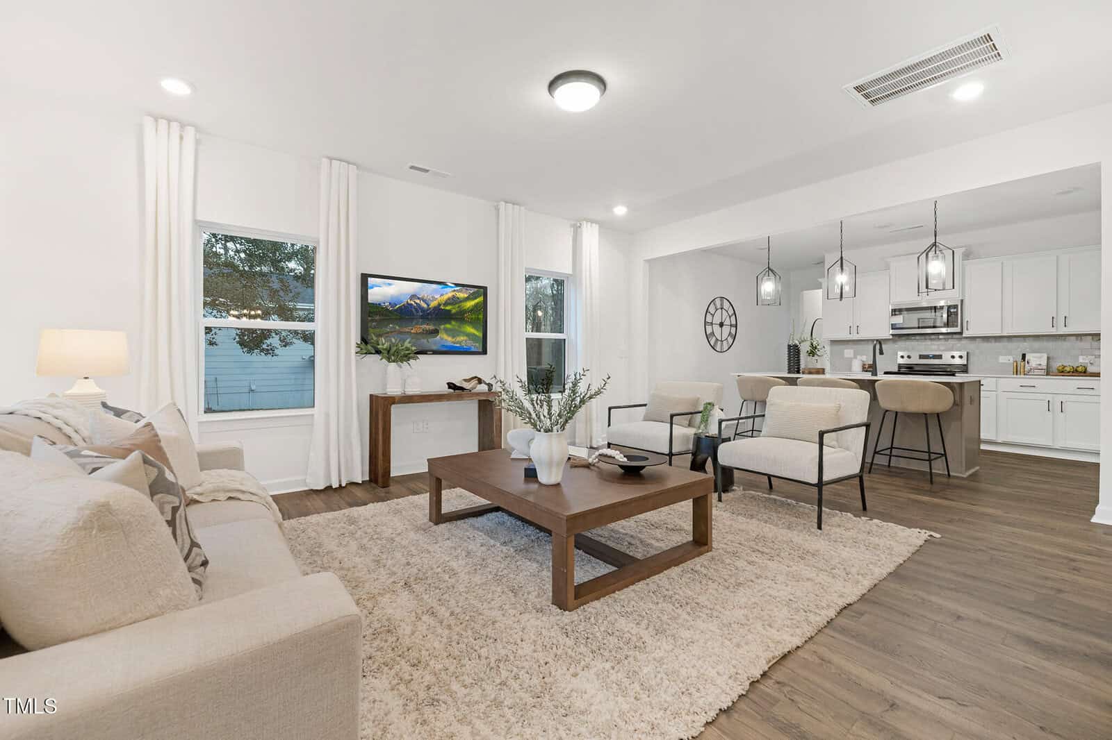 Modern open-concept living room and kitchen with neutral tones, beige sofa, armchairs, wooden coffee table, rug, TV, and kitchen island with bar stools under pendant lights.