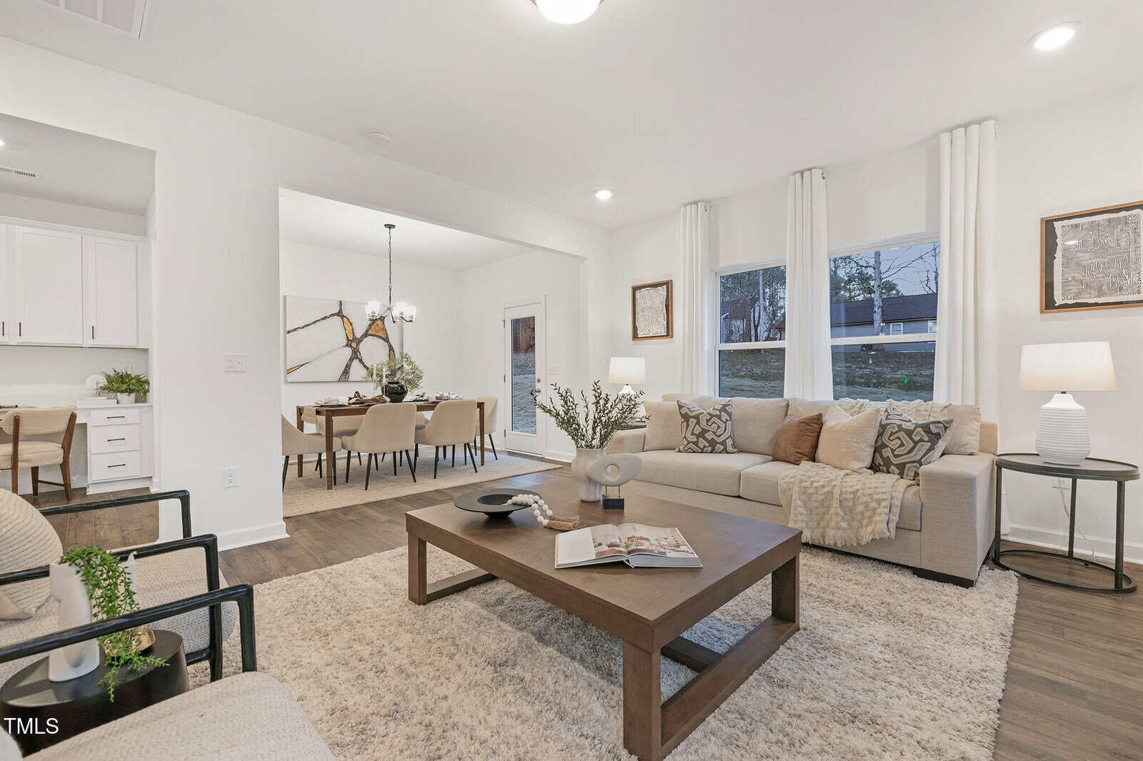 Modern living room with beige sofas, a wooden coffee table, and a soft rug. The space opens to a dining area with a long table, chairs, and a chandelier. Large windows bring in natural light, and the decor is neutral and cozy.