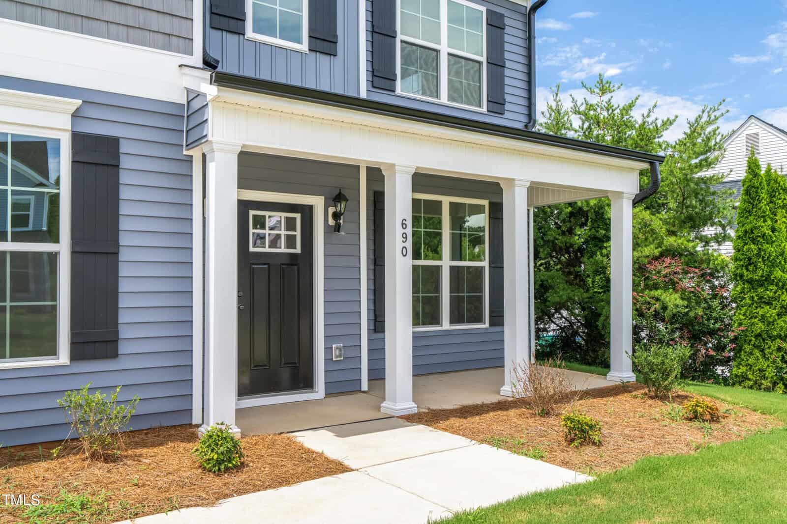 A two-story house with blue siding, white trim, and black shutters. The front door is black, beneath a covered porch supported by four white columns. The house number 690 is visible on one column. Shrubs line the walkway.