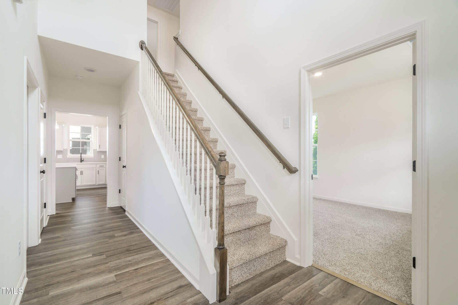 A bright hallway with wood-look flooring, a carpeted staircase with a wooden handrail, an open doorway to a carpeted room on the right, and a view into a white kitchen at the end of the hall.