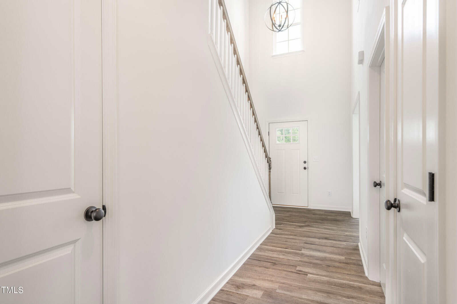 Bright entryway with light-colored walls, wood-look flooring, a staircase on the left with a dark banister, a white front door with glass panels, and a tall window above the door letting in natural light.