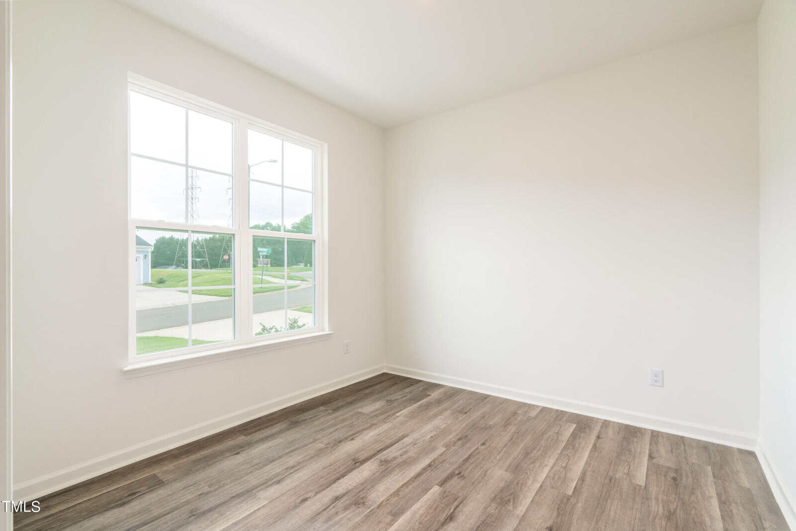 An empty room with light-colored walls, a large window letting in natural light, and wood-look vinyl flooring. The window offers a view of greenery and a street outside.