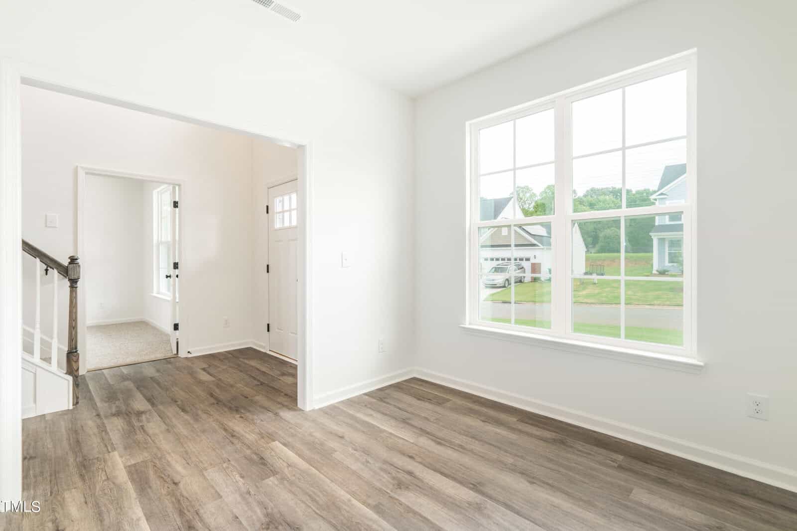 A bright, empty room with light-colored walls, large windows, and wood-style flooring. There is a view of a staircase, another room, and an entrance door. Houses and greenery are visible outside the window.