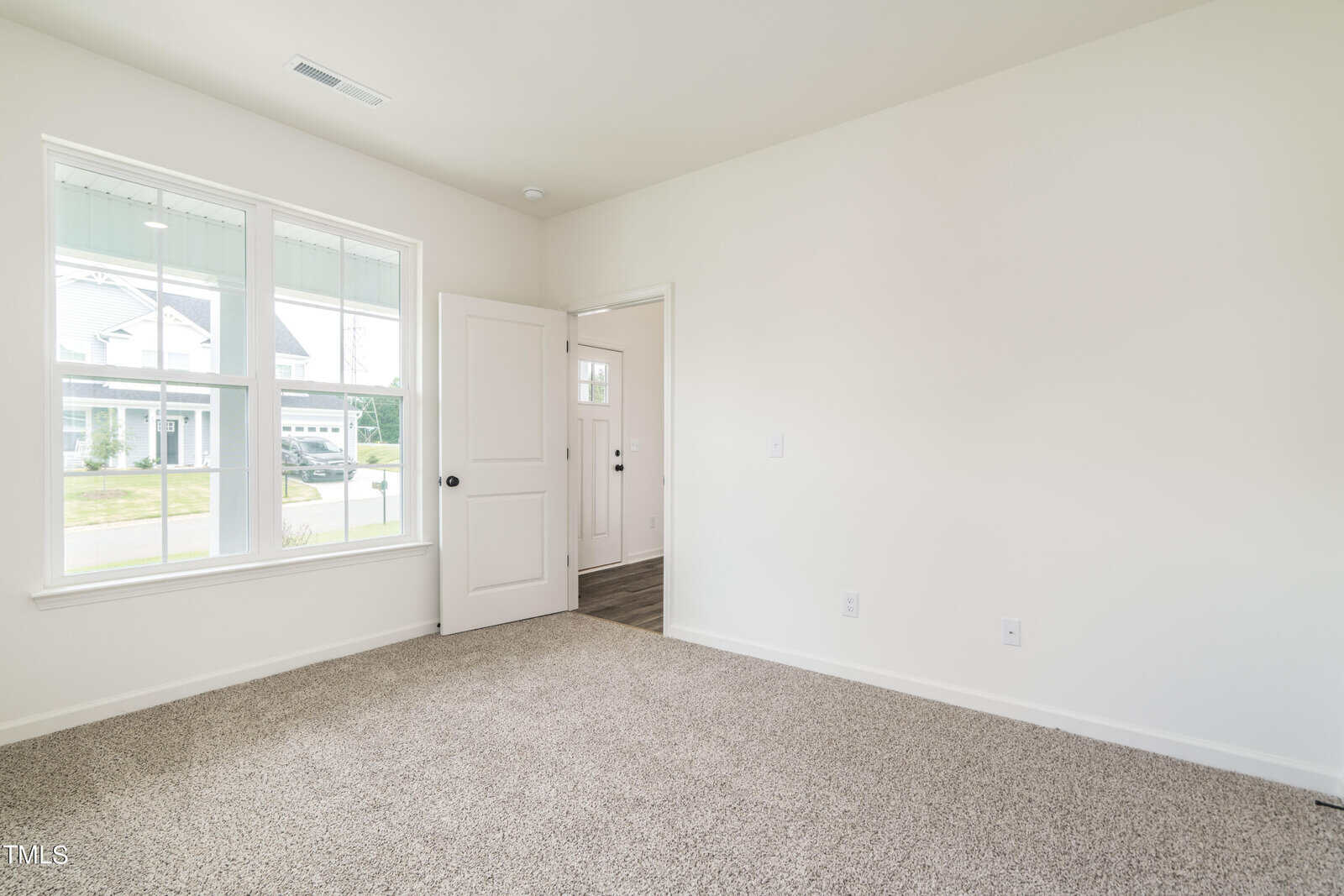A bright, empty room with beige carpet, white walls, a large window showing a suburban street, and a white door leading to a hallway with wood flooring.