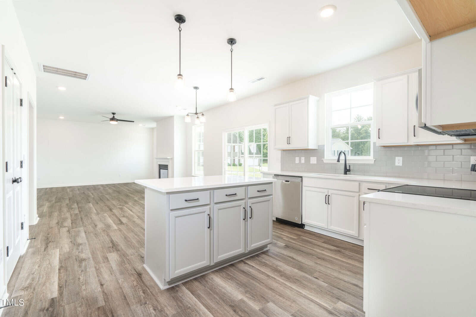 Bright, modern kitchen with white cabinets, gray island, stainless steel appliances, pendant lighting, and wood-look flooring, open to a spacious living area with a ceiling fan and fireplace. Large windows let in natural light.