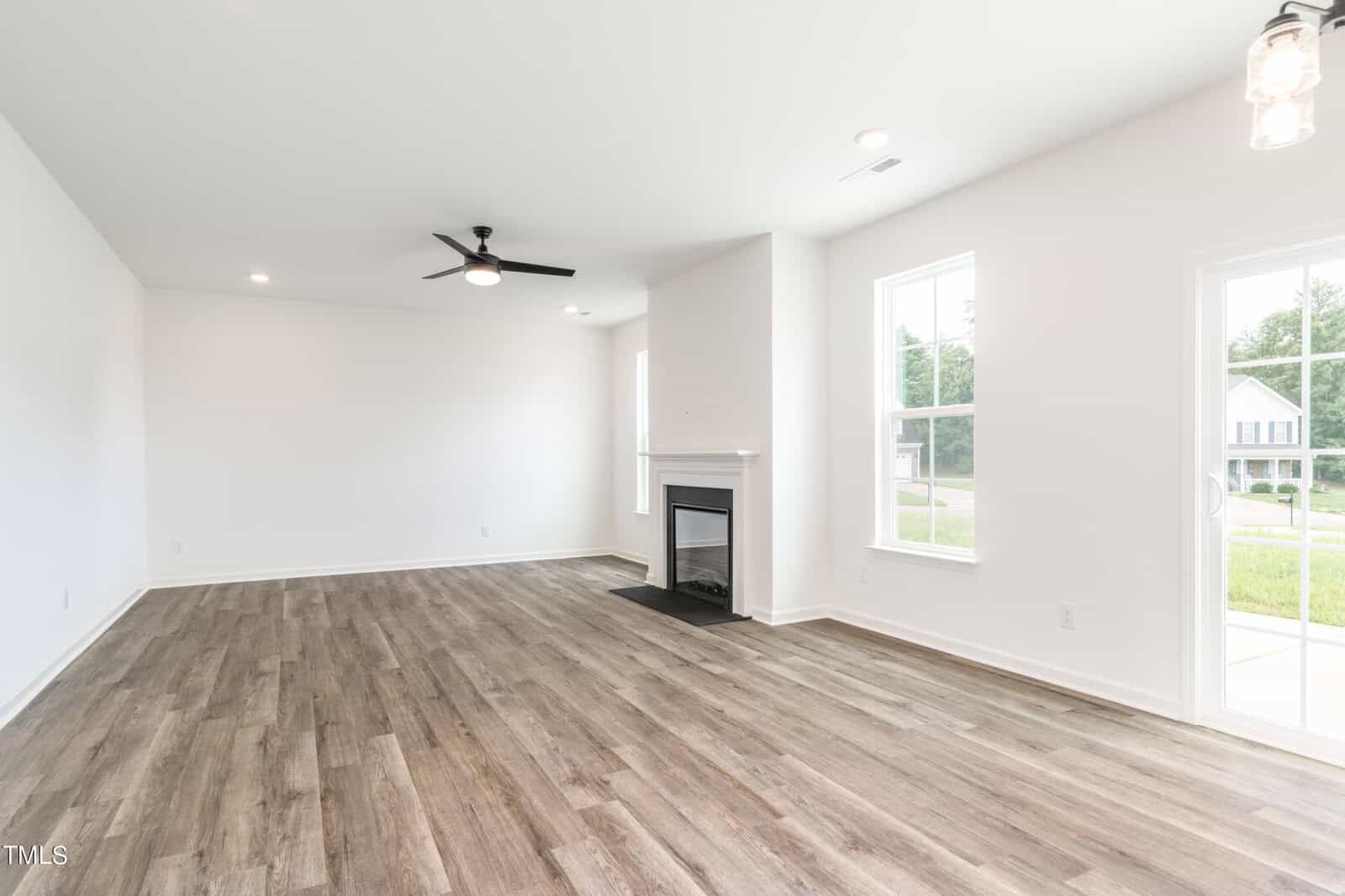 A spacious, empty living room with light-colored walls, wood-style flooring, a ceiling fan, a fireplace, large windows, and a glass door leading outside. The room is bright with natural light.