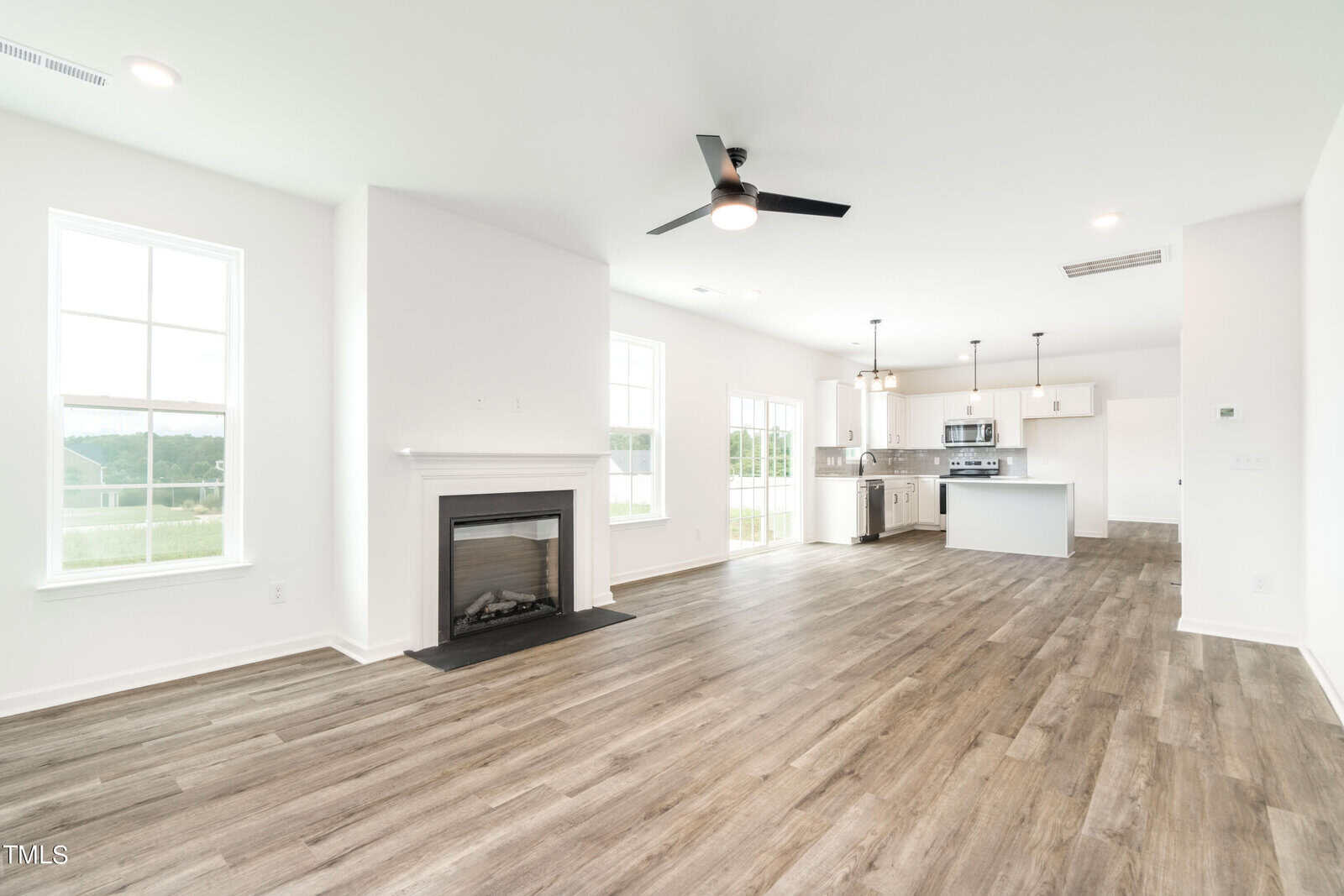 A spacious, unfurnished open-plan living area with light wood flooring, white walls, a black ceiling fan, a fireplace, large windows, and a modern kitchen with white cabinets and stainless steel appliances in the background.