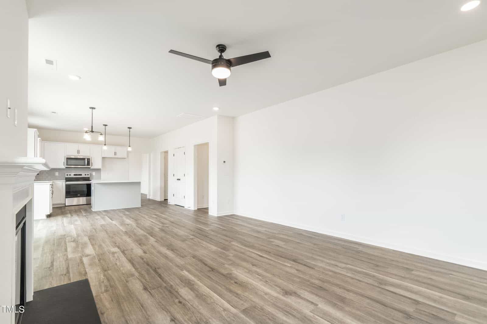 Spacious, modern living area with wood flooring, white walls, a ceiling fan, and an open kitchen featuring stainless steel appliances and an island.
