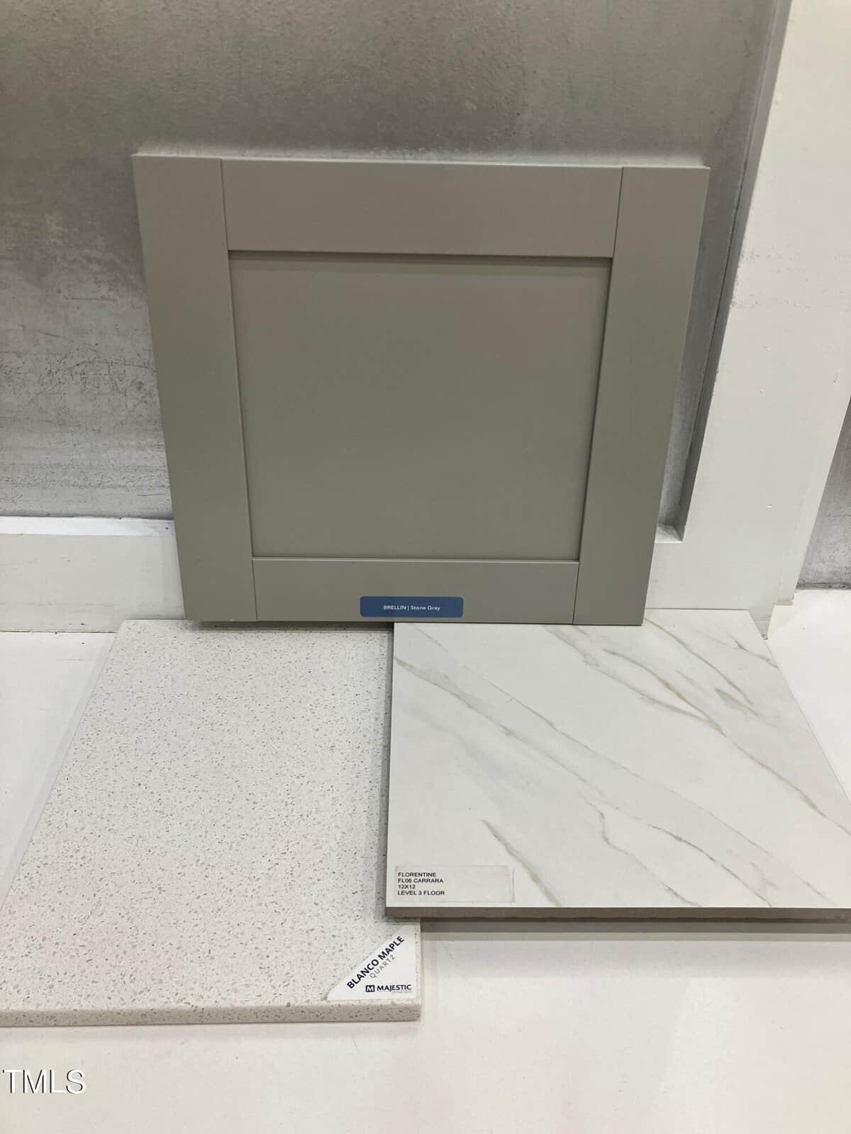 A gray cabinet door sample stands behind two countertop samples: one speckled white and one white with subtle gray veining, all displayed on a white surface against a light gray wall.