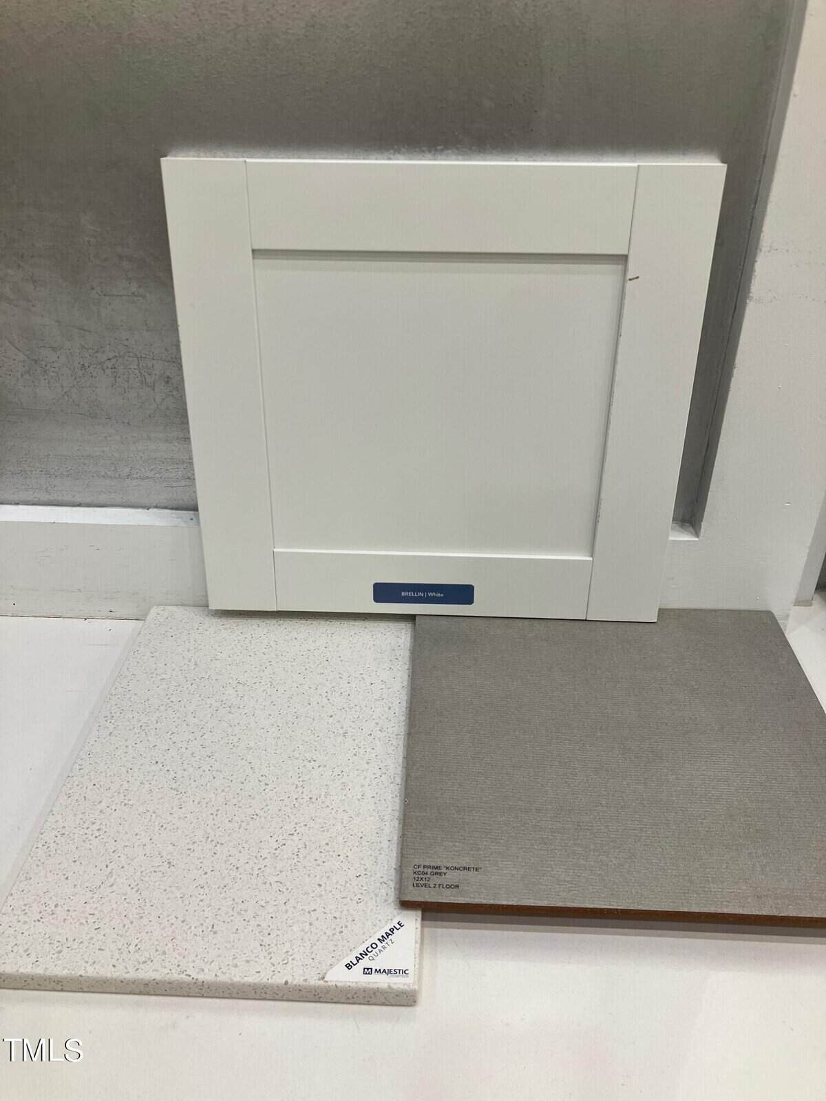 A white cabinet door sample stands upright behind two countertop samples, one with a white speckled surface and the other a solid gray color, all displayed on a white surface against a gray and white background.