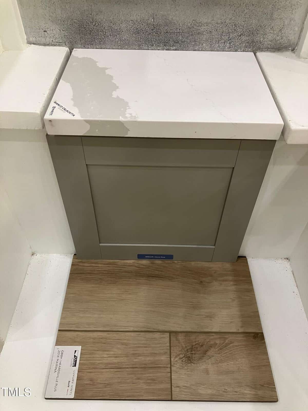 A sample display shows a white countertop, a gray cabinet door, and light wood-look floor tiles arranged vertically, each with a small label attached. The display is surrounded by white walls on three sides.