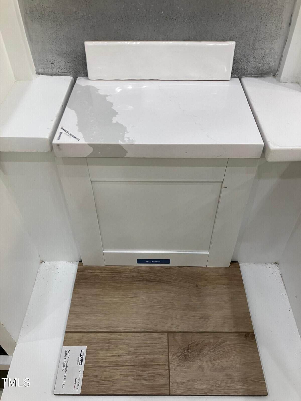 A white cabinet with a marble-patterned countertop and a wooden floor sample in front. A small backsplash is installed above the counter, and there is a label on the floor sample.