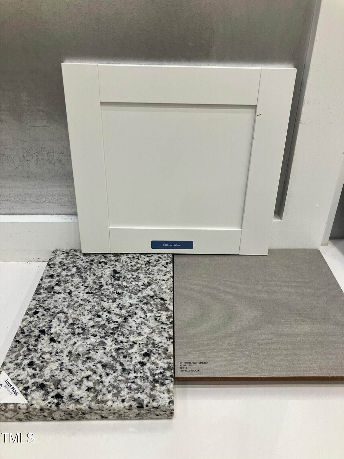 A white cabinet door sample stands upright behind a speckled gray granite countertop sample and a large square beige flooring tile sample, which are laid flat on a white surface.