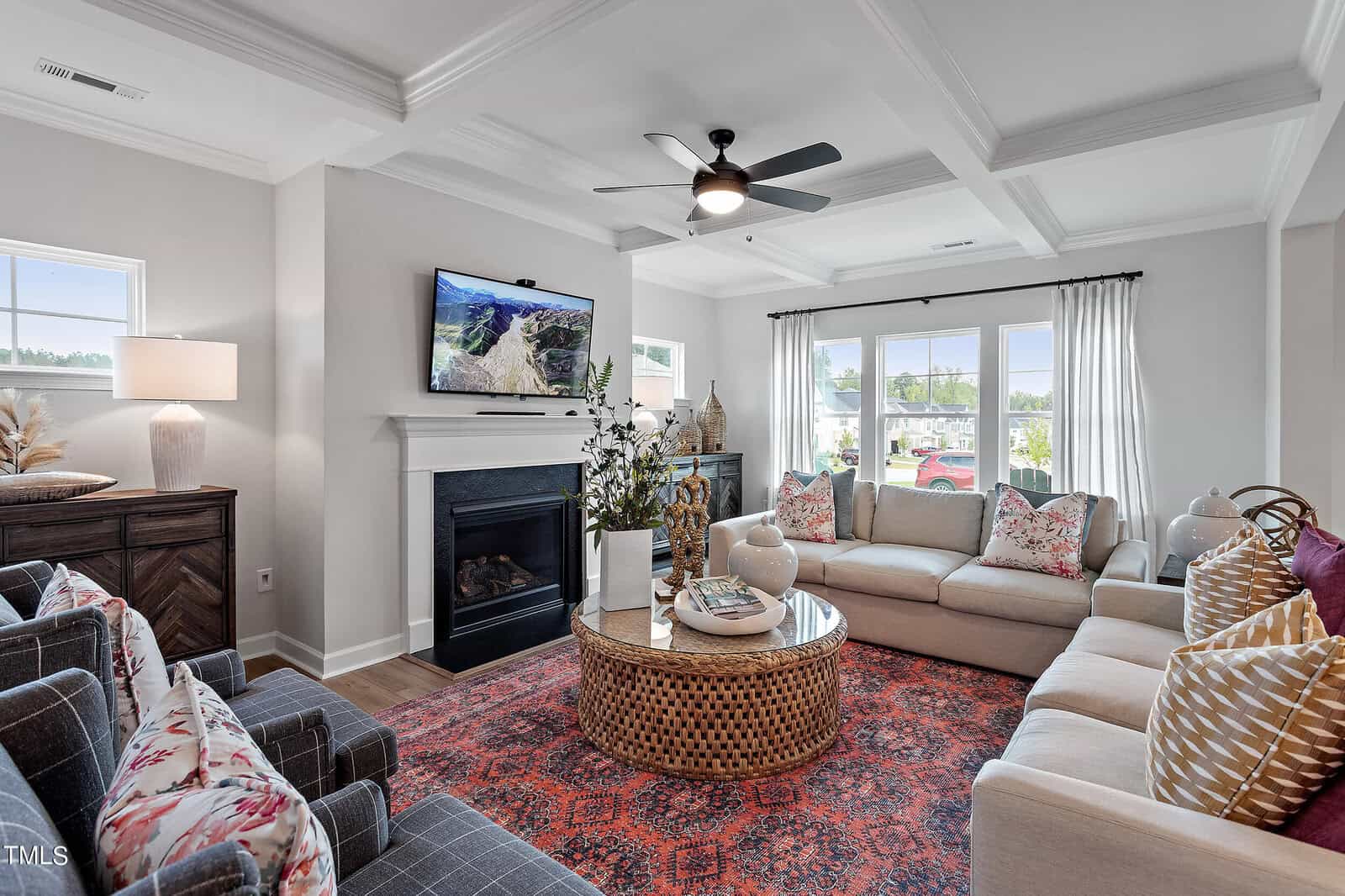 A bright living room with two beige sofas, patterned pillows, a round wicker coffee table, a red rug, a fireplace with a TV above it, large windows with white curtains, and decorative accents on cabinets.