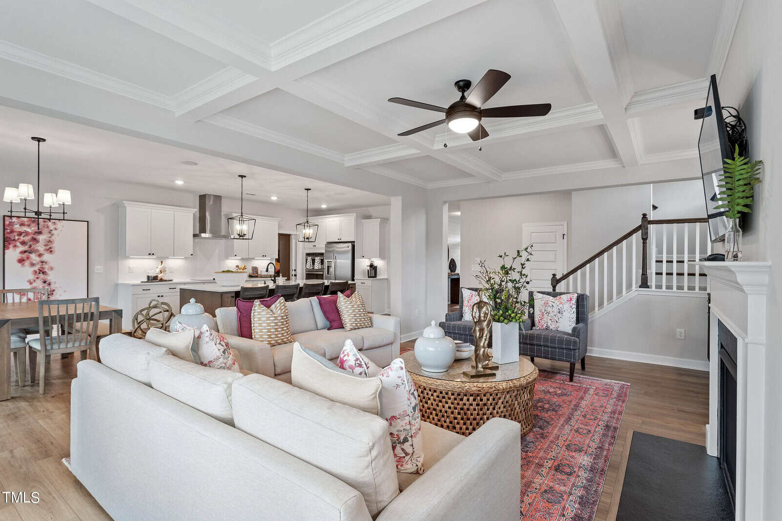 Open-concept living area with two beige sofas, colorful throw pillows, a round coffee table, and a TV mounted above a fireplace. The kitchen, dining area, and staircase are visible in the background.