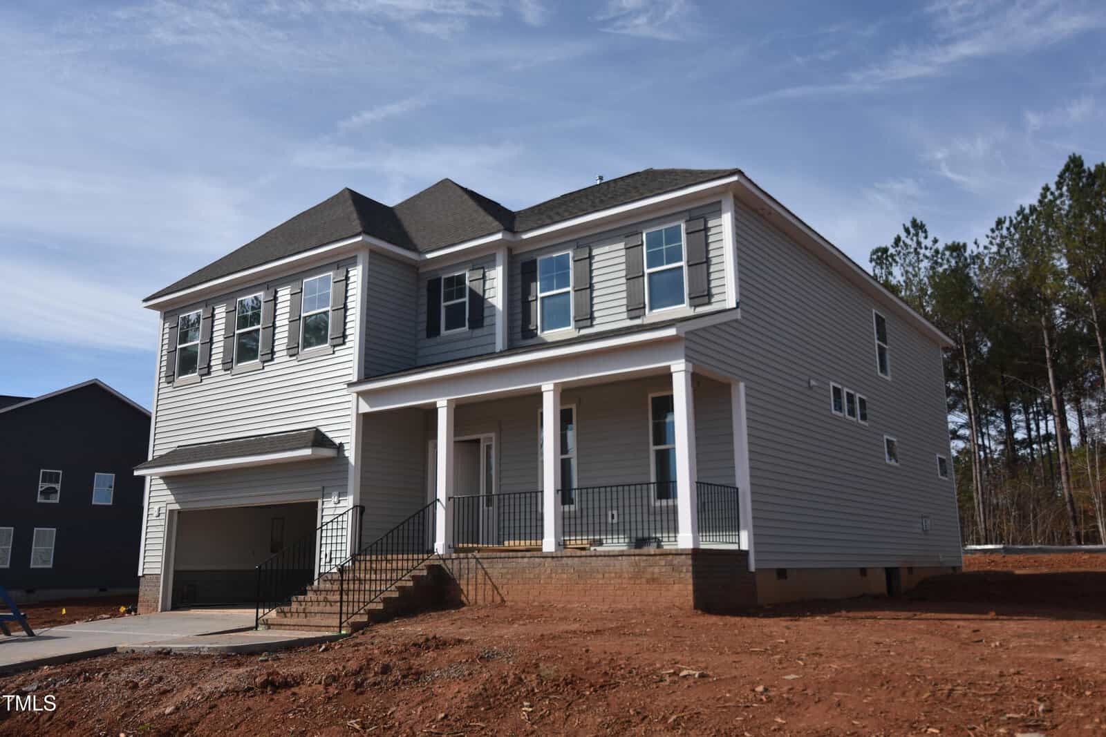 Two-story gray house with white trim and a front porch, featuring a double garage and several windows. The yard is unfinished with bare soil, and there are trees and another house in the background.