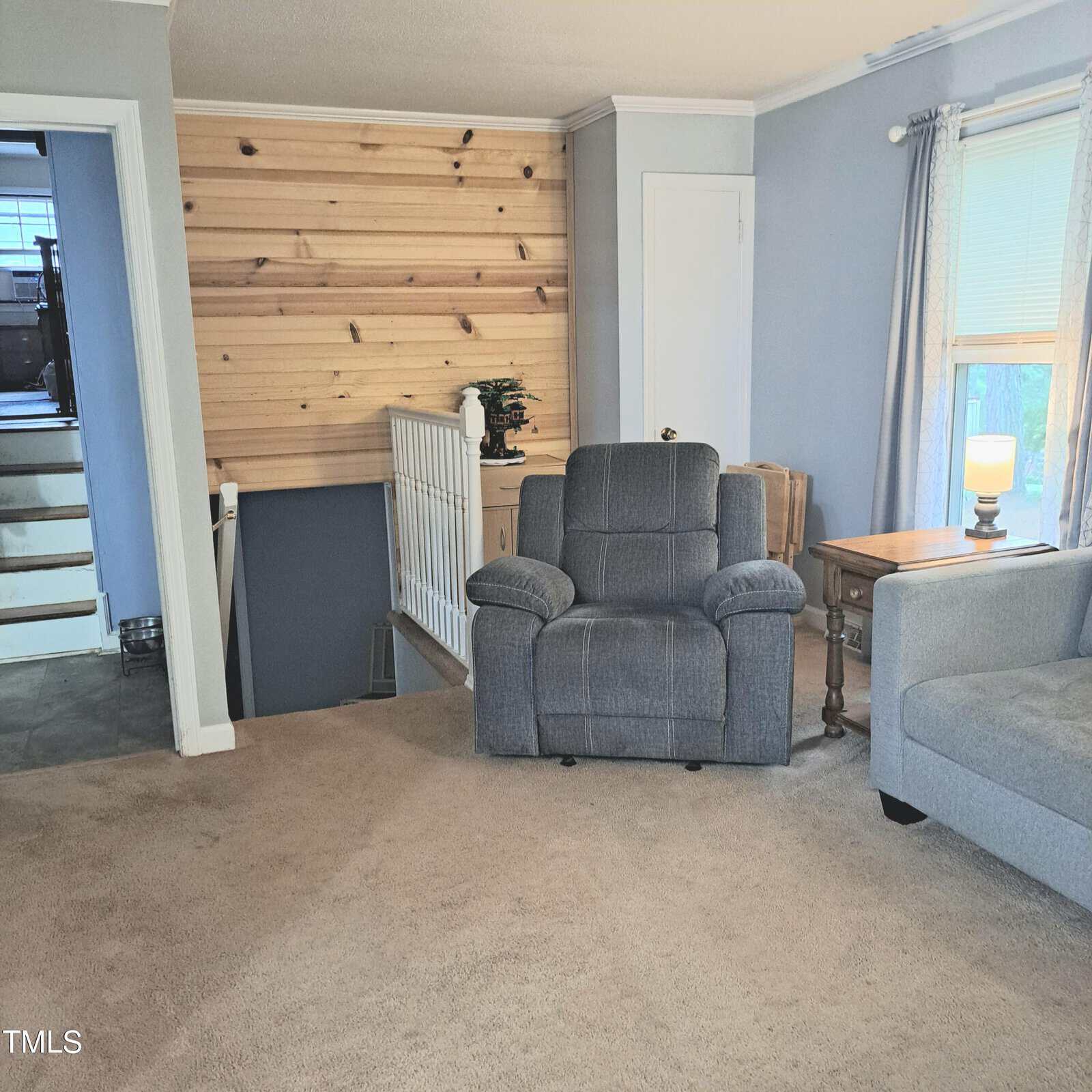 A living room with a gray recliner, a gray sofa, and a wooden side table with a lamp. Light blue walls and a natural wood accent wall are visible. A staircase with a white railing leads downstairs. A window with sheer curtains lets in sunlight.