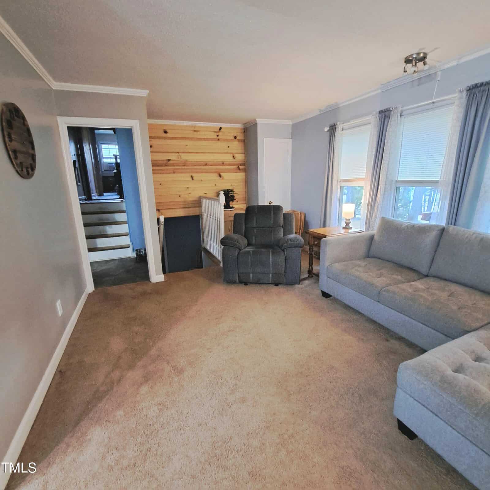 A cozy living room with gray walls and carpet, featuring a gray sectional sofa and a recliner. Large windows with curtains allow natural light. A wooden accent wall is visible near the steps leading to another room. A round clock decorates the wall.