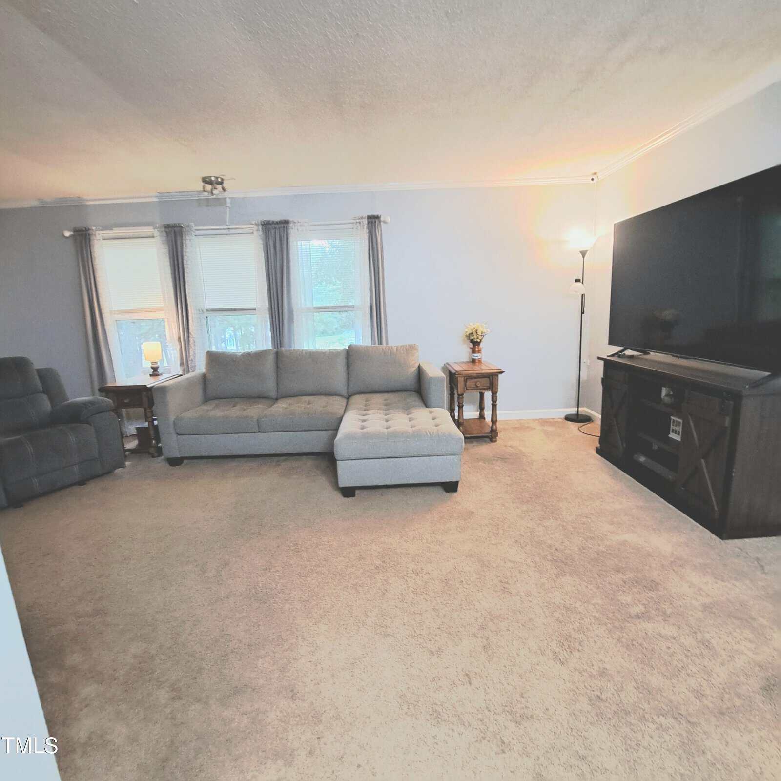 A living room featuring a gray sectional sofa with an ottoman, a large flat-screen TV on a wooden stand, a recliner chair, and a floor lamp. The room has light-colored carpet and two windows with gray curtains. A small side table holds a vase of flowers.