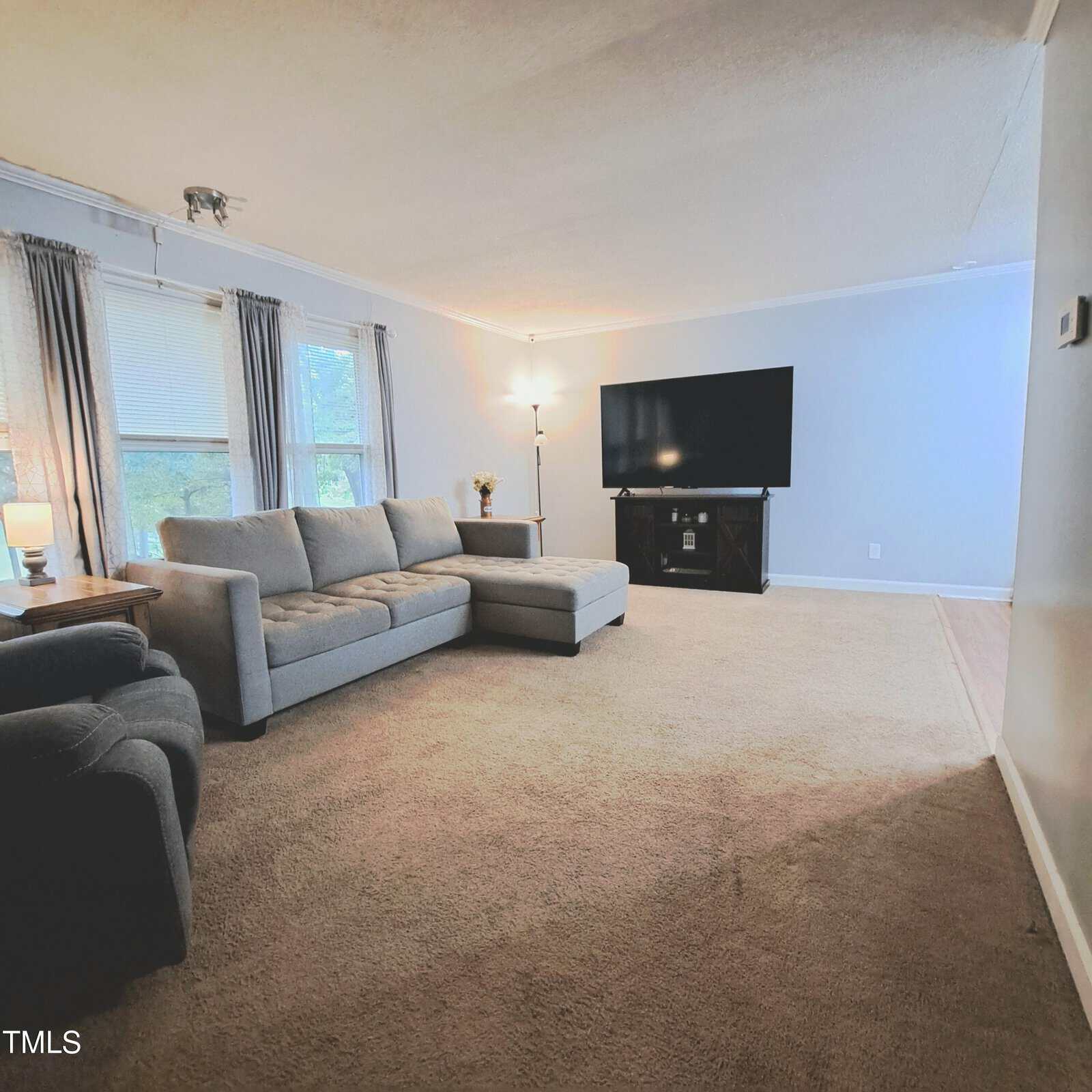 A spacious living room with a gray sectional sofa and an armchair on a beige carpeted floor. A large flat-screen TV is positioned on a stand against the wall. Two windows with curtains allow natural light to brighten the room.